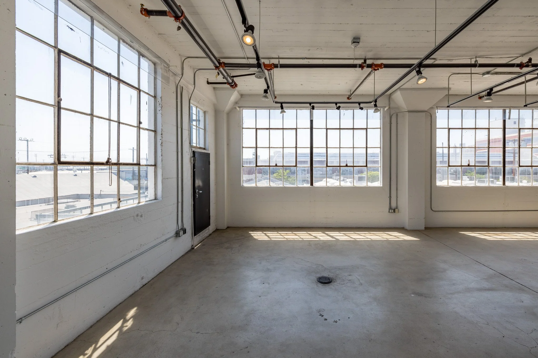 Empty industrial space with large windows, exposed pipes, and concrete floor.