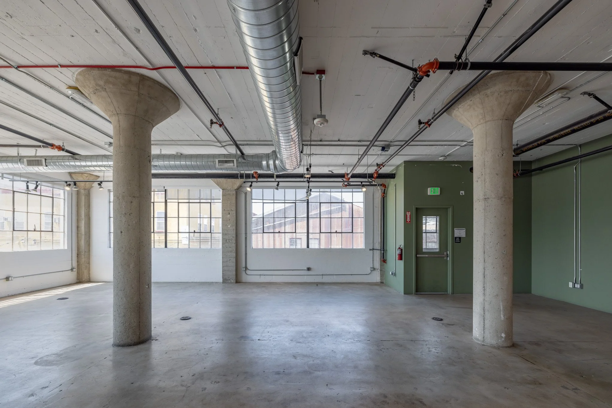 Empty industrial-style interior with concrete columns, large windows, exposed ductwork, and a green wall with an exit door.