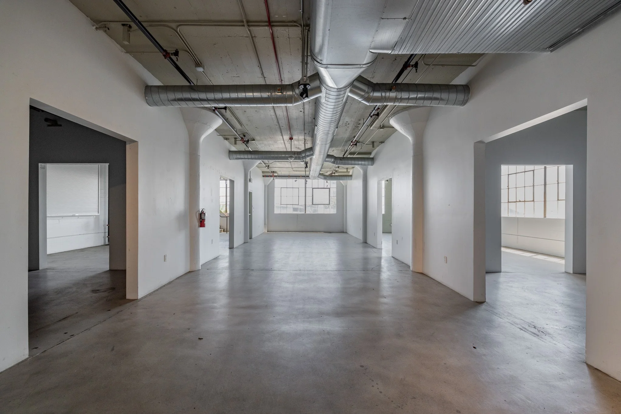 Empty industrial-style office space with white walls, large windows, exposed ductwork and concrete floors.