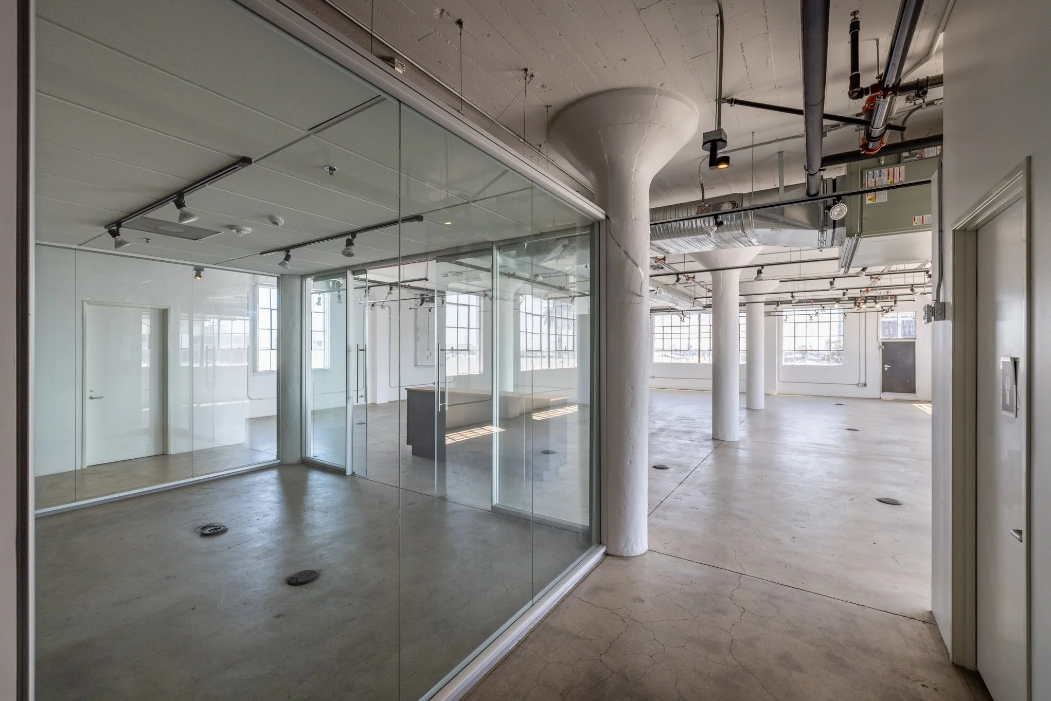 Empty industrial-style office space with glass walls, large windows, white columns, and exposed ductwork.