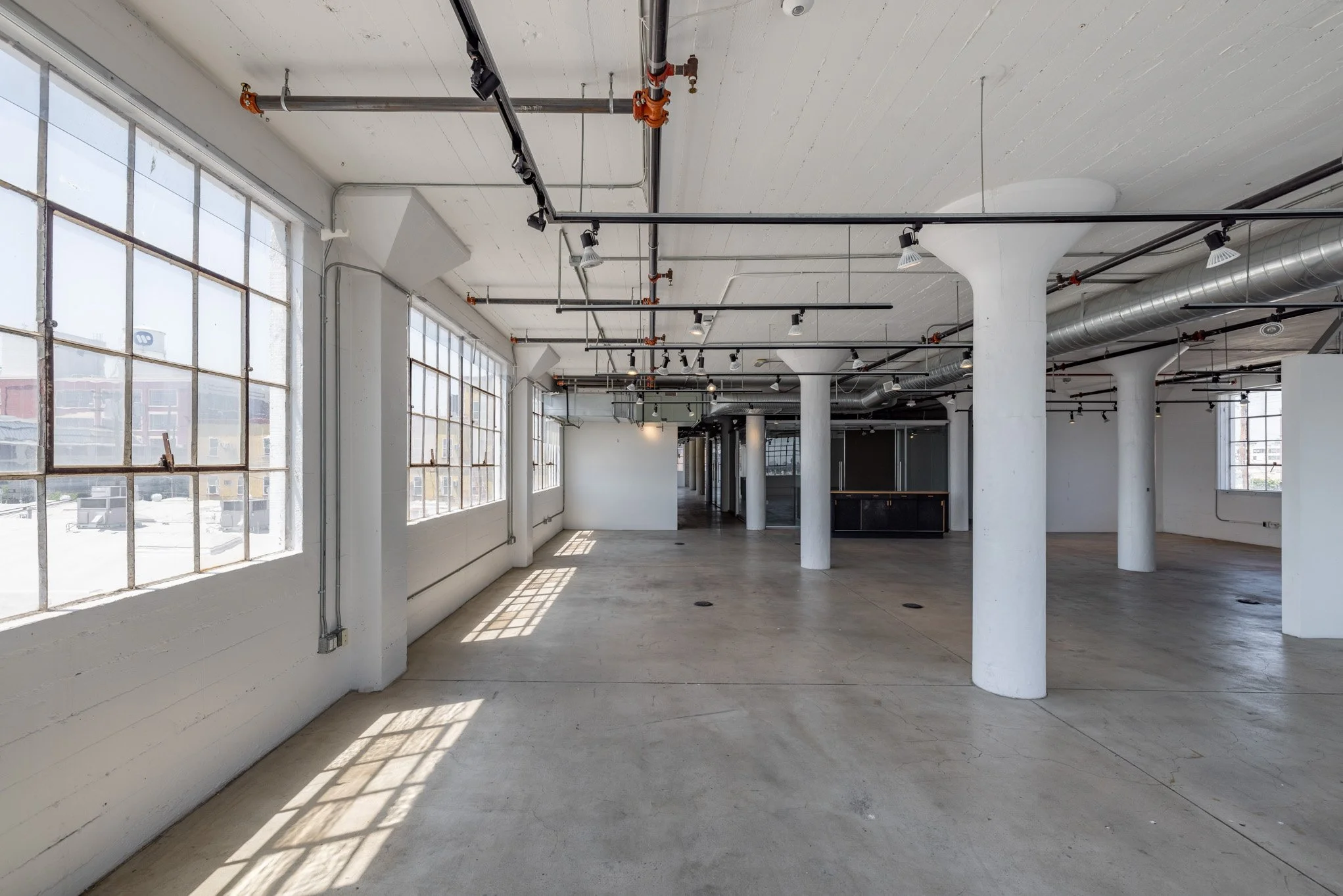 Empty industrial-style loft with large windows, white walls, concrete floors, and exposed pipes and columns.