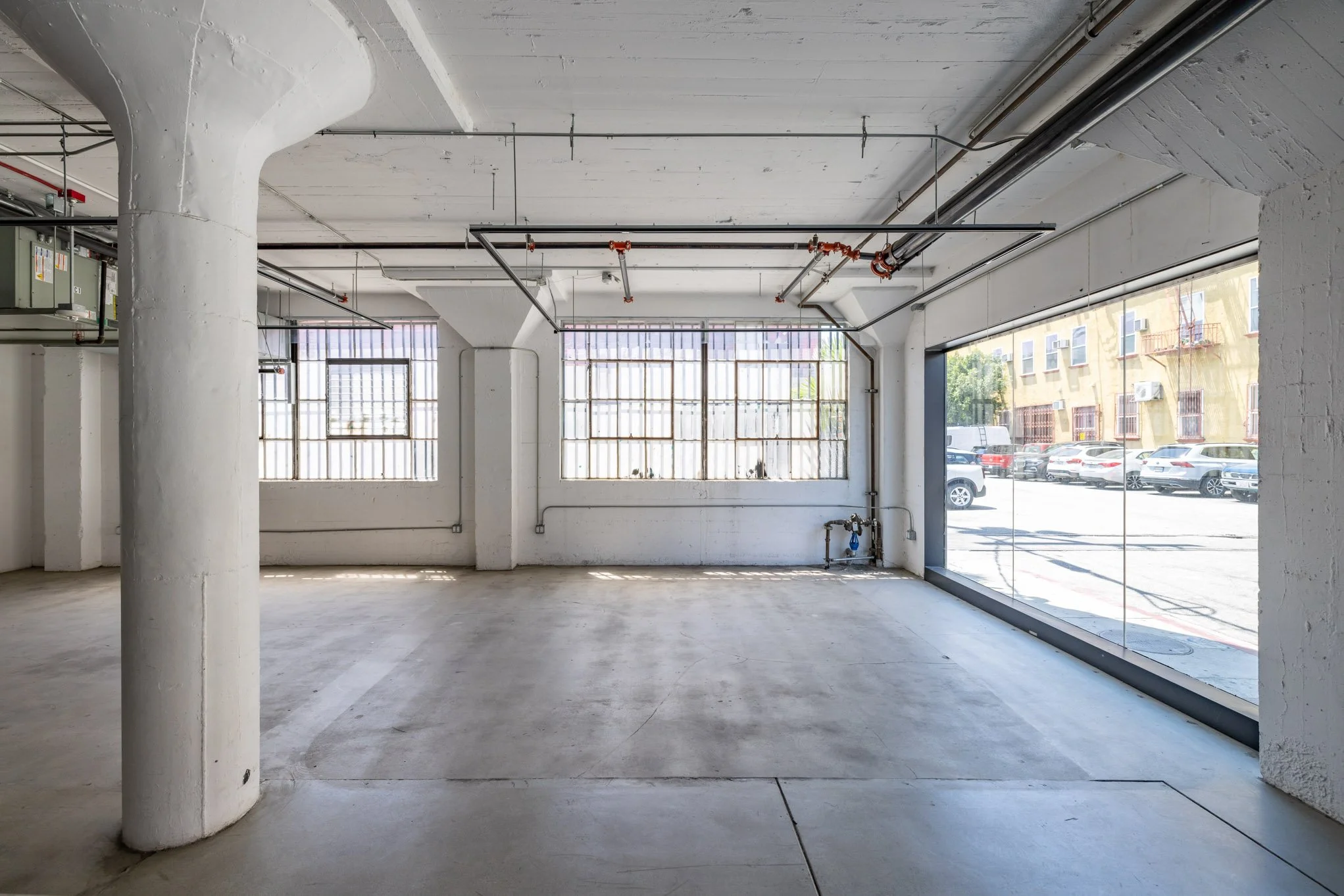 Empty commercial space with large front windows, white walls and ceiling, concrete floor, and visible piping and electrical fixtures.