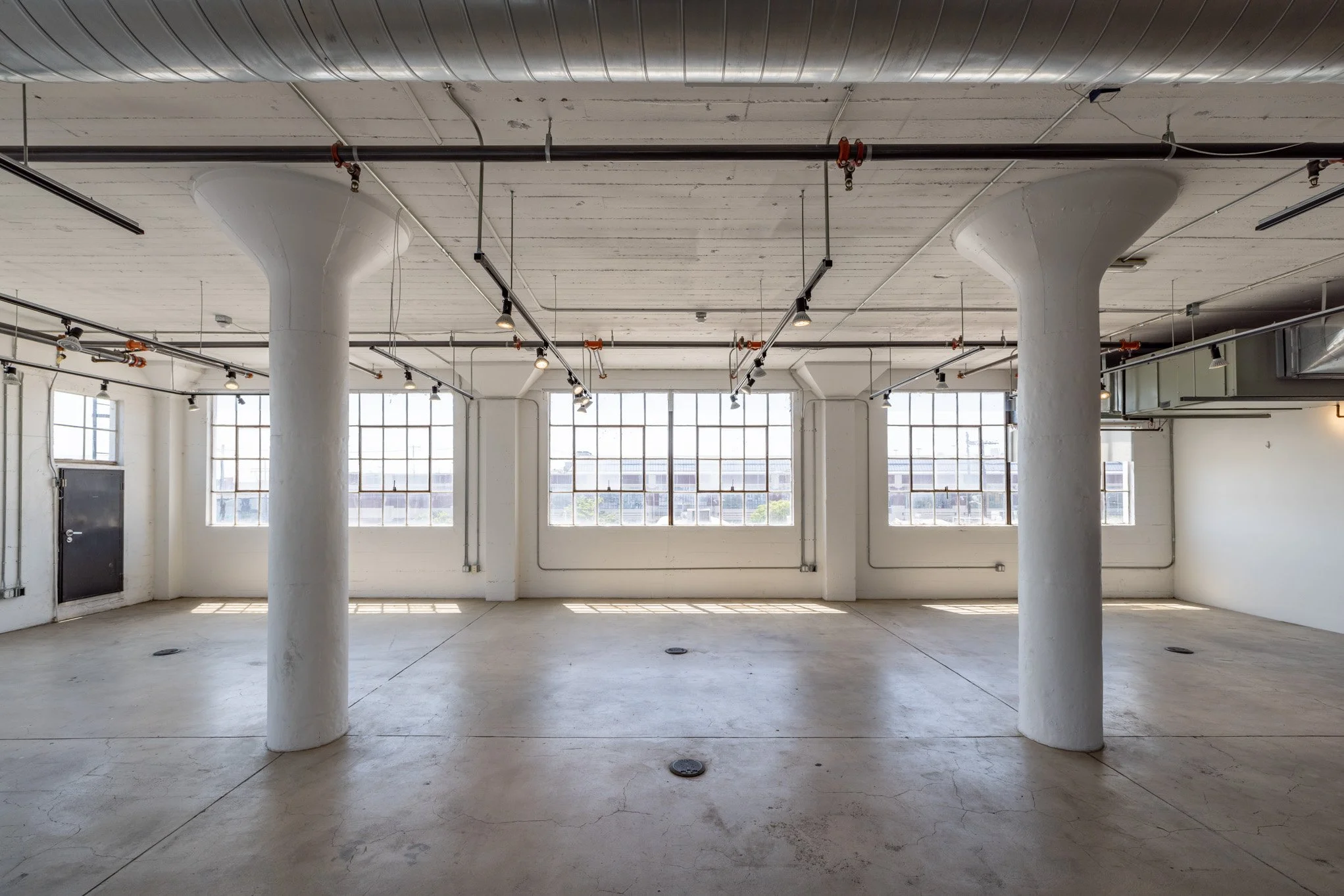 Empty industrial-style loft with large windows, white walls, exposed ceiling pipes, and concrete floor.