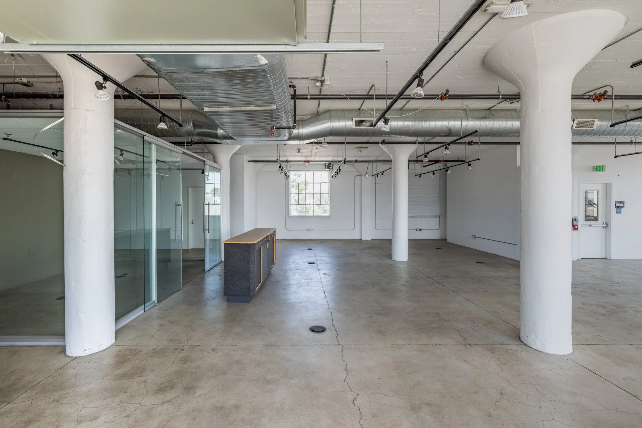 Empty commercial space with concrete floors, white walls, large windows, visible pipes and ducts on the ceiling, and a glass partition on the left side.