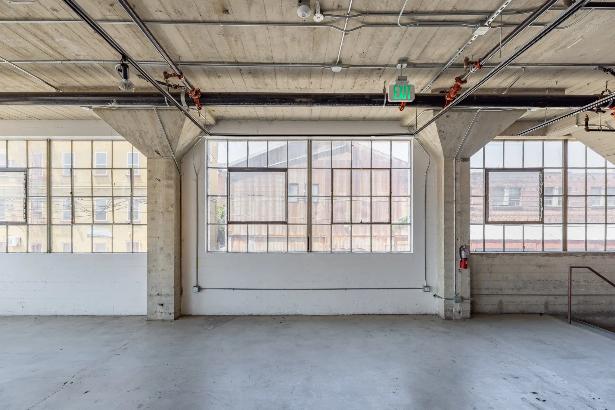 Empty industrial-style room with large windows, concrete flooring, exposed pipes, and an emergency exit sign.