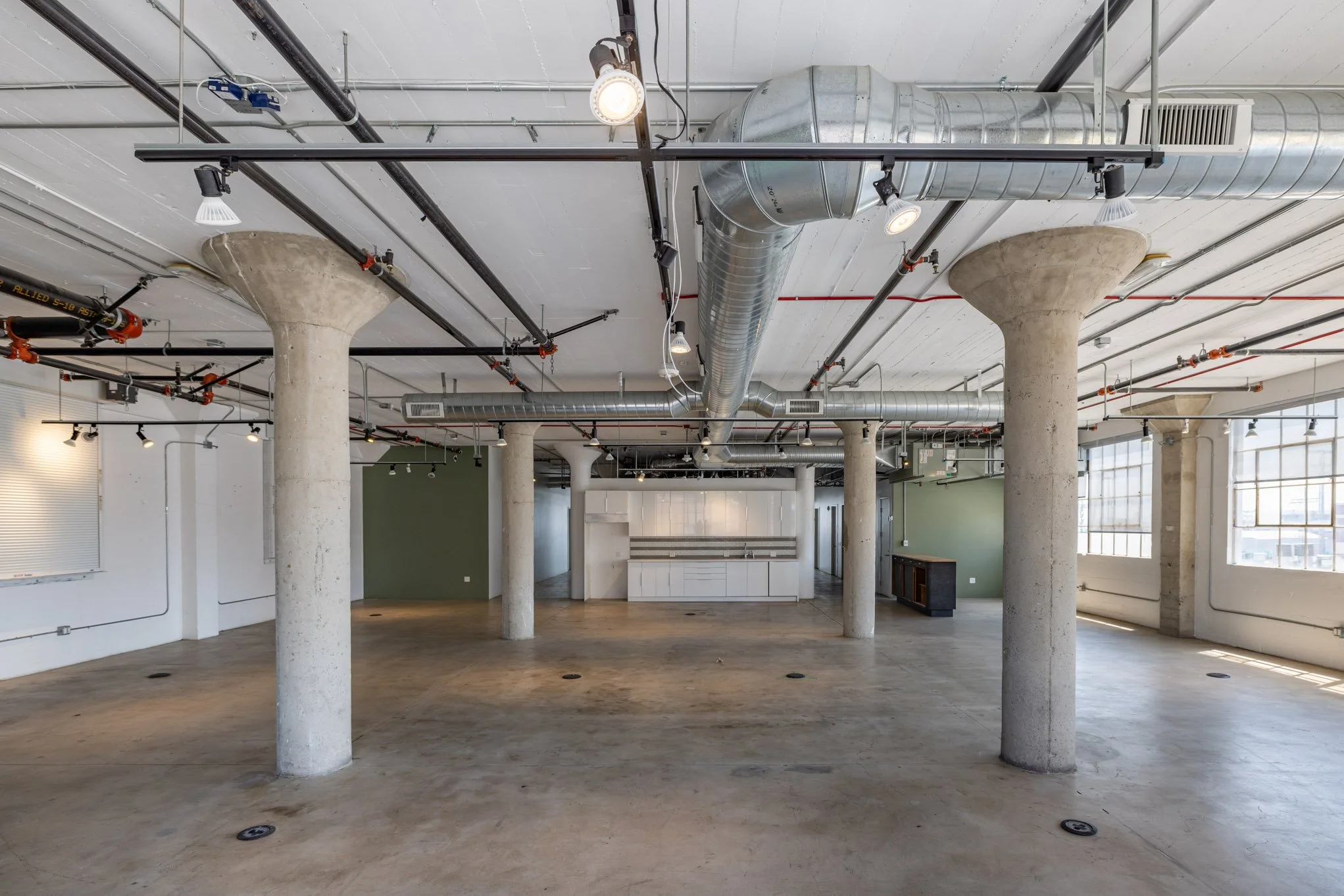 Empty industrial-style loft with exposed ductwork, concrete columns, and large windows.