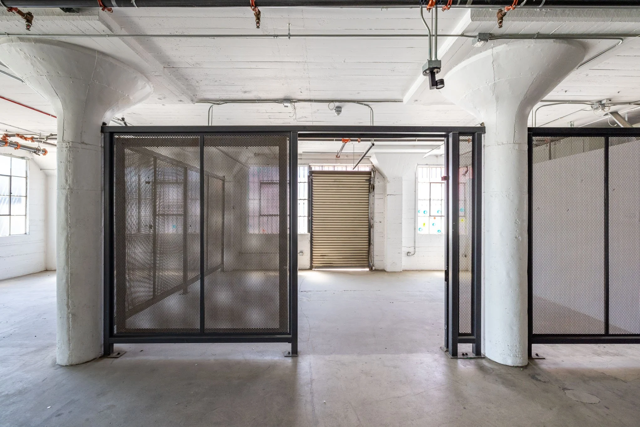 An interior view of an industrial space with white walls, large windows, exposed pipes, and a metal gate with perforated panels.