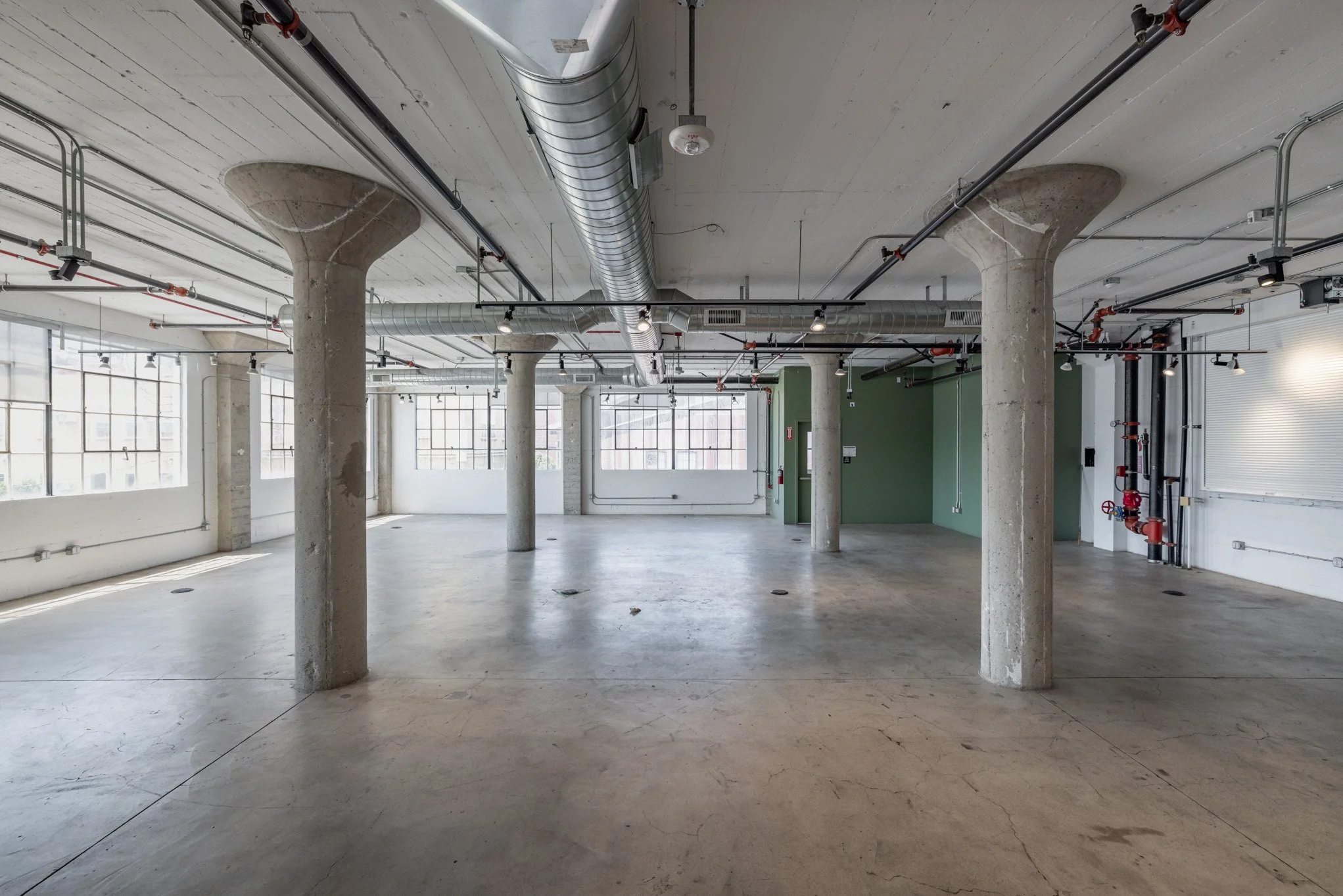 Empty industrial-style room with concrete pillars, large windows, exposed ductwork, and pipes.
