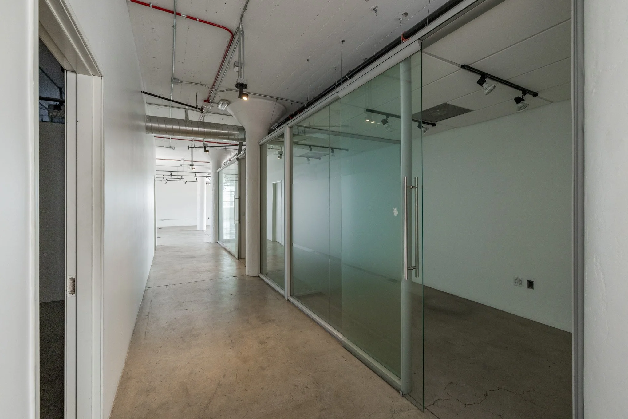 Empty modern office corridor with glass-walled rooms, white walls, and exposed ceiling pipes and ducts.