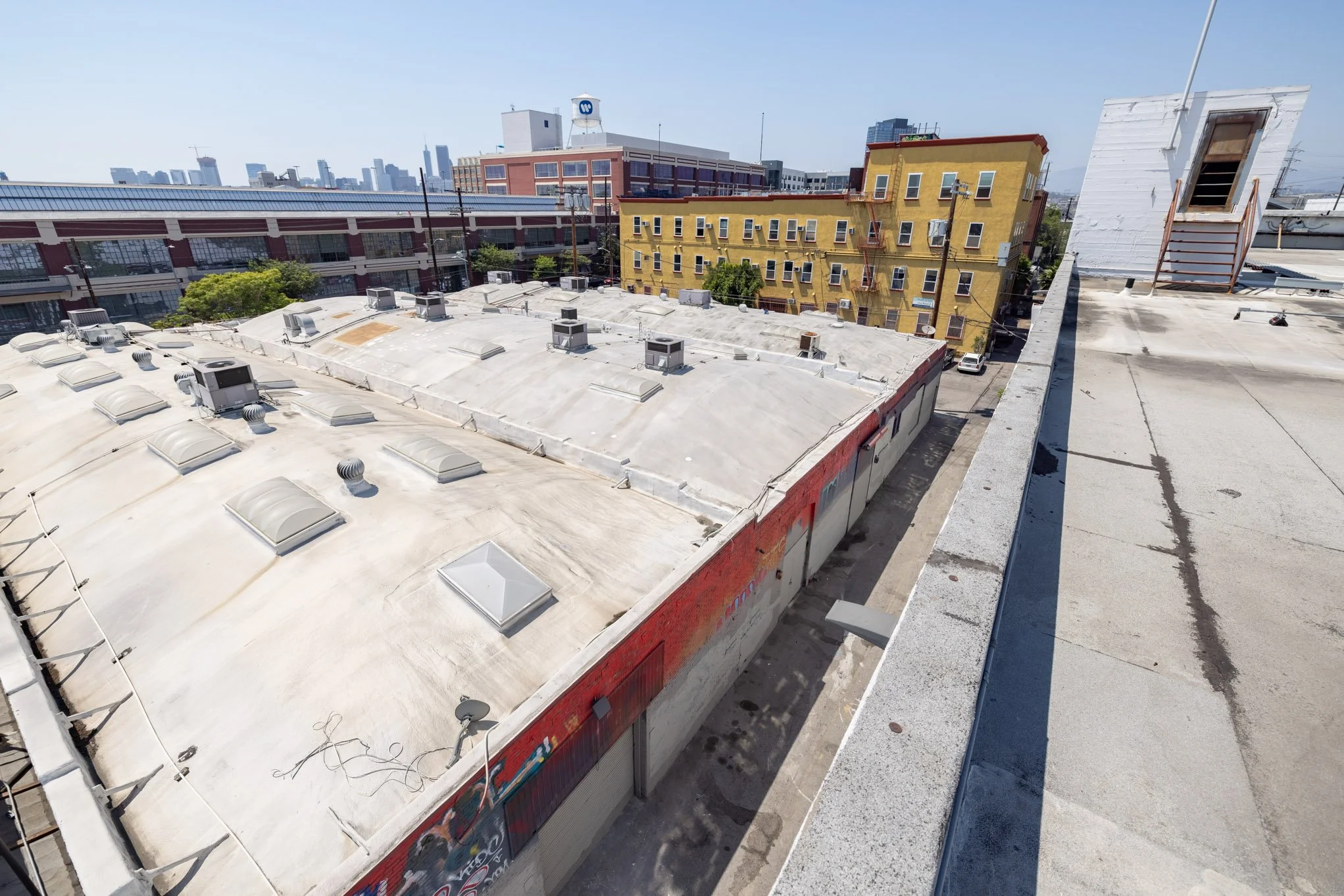View of city rooftops, including a large flat roof with multiple vents and HVAC units, and a yellow building across the street with a skyline in the background.