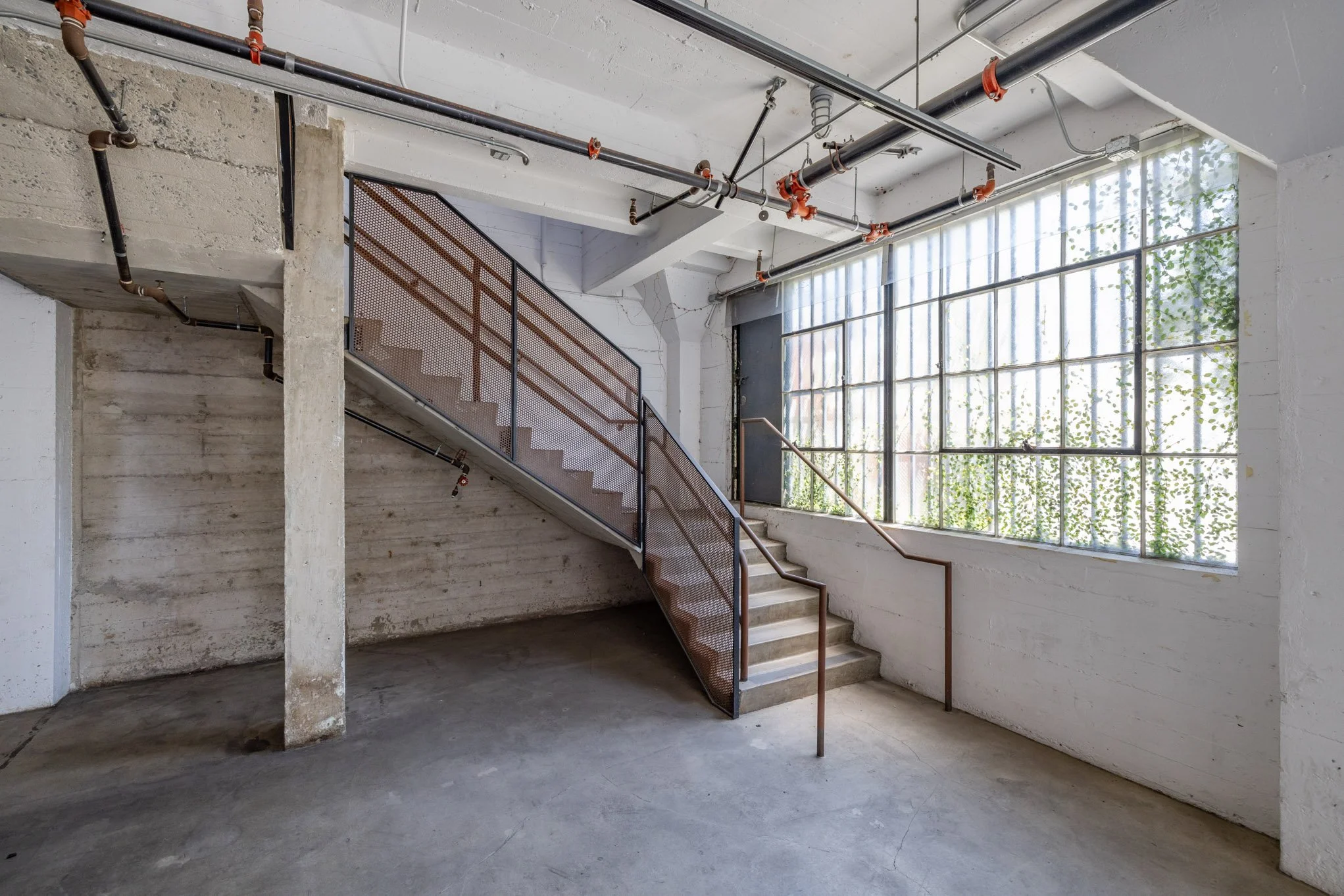 Interior of an industrial-style building with concrete walls, a staircase with a metal railing, large grid-style windows, and exposed ceiling pipes.