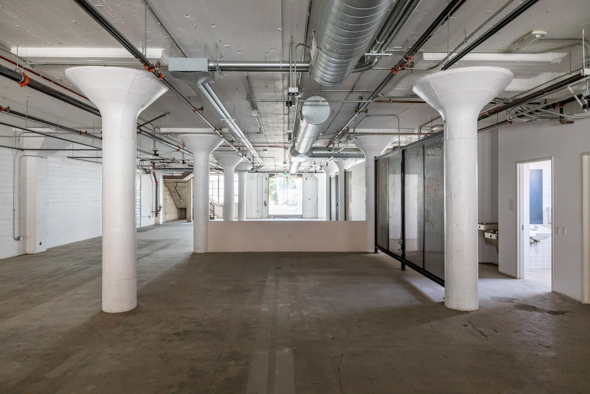Empty industrial-style space with white columns, exposed ceiling pipes, and a small bathroom with a sink on the right.