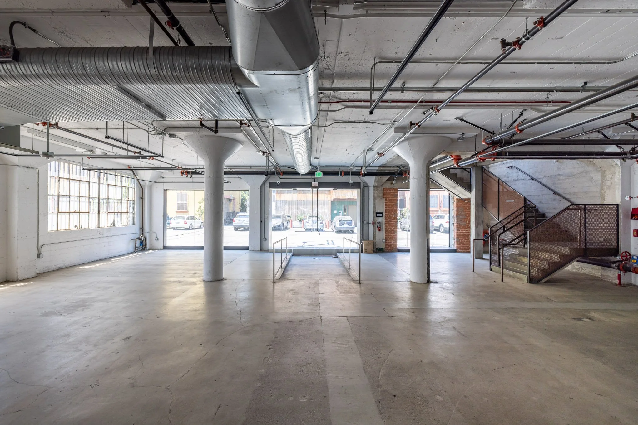 Empty industrial-style commercial space with large glass windows, concrete floors, exposed piping and staircase, and no furniture.