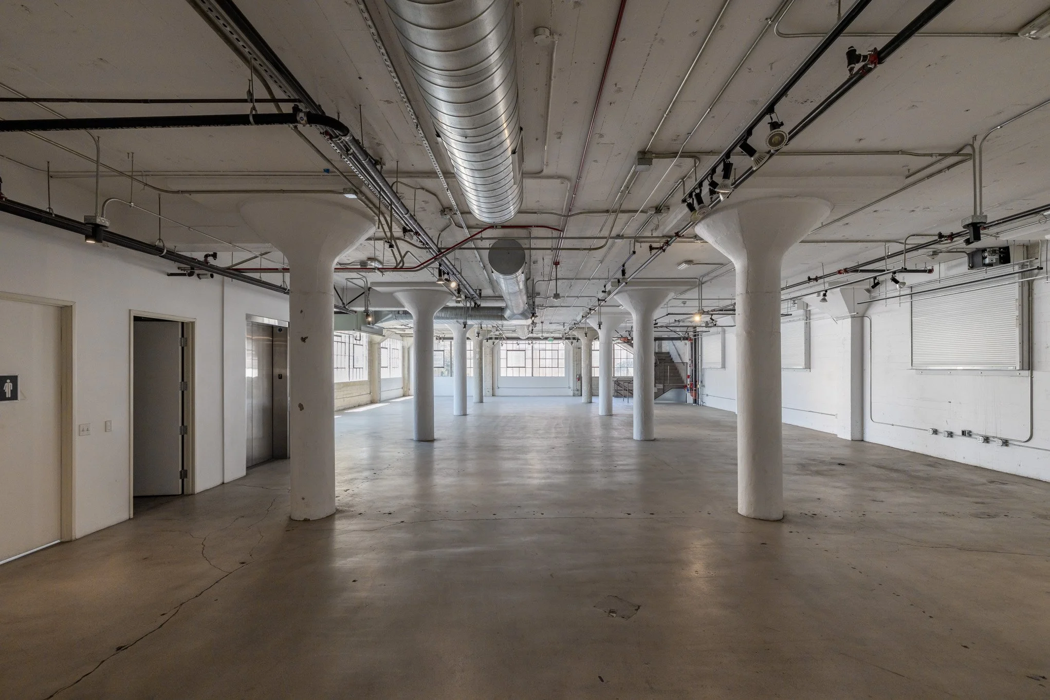 Empty industrial space with white pillars, large windows, and exposed ceiling pipes and ductwork.