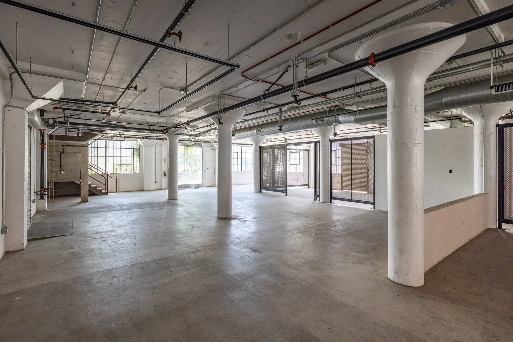 Empty industrial or commercial space with concrete floors, large windows, white columns, and exposed ceiling pipes.