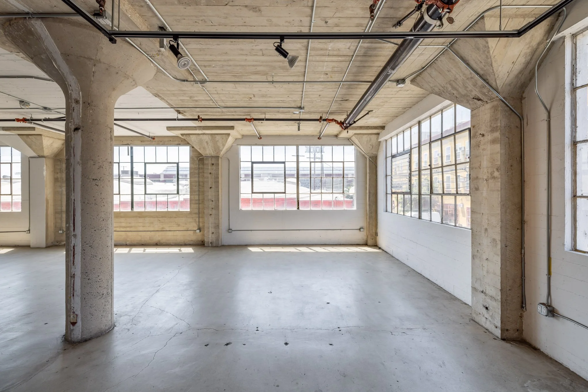 Empty industrial-style loft with large windows, exposed concrete columns, ceiling pipes, and a concrete floor.