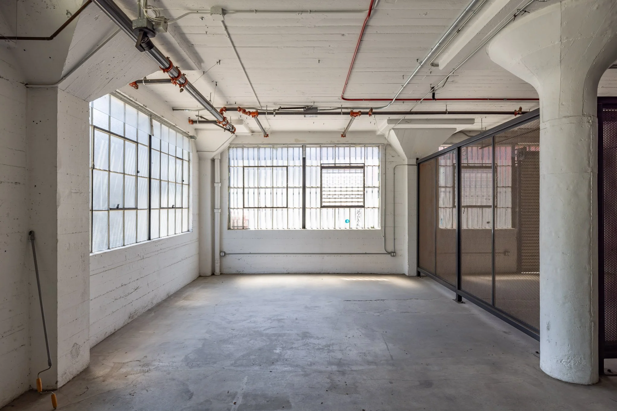 Empty industrial-style room with large windows, exposed pipes, a partitioned area, and concrete floors.