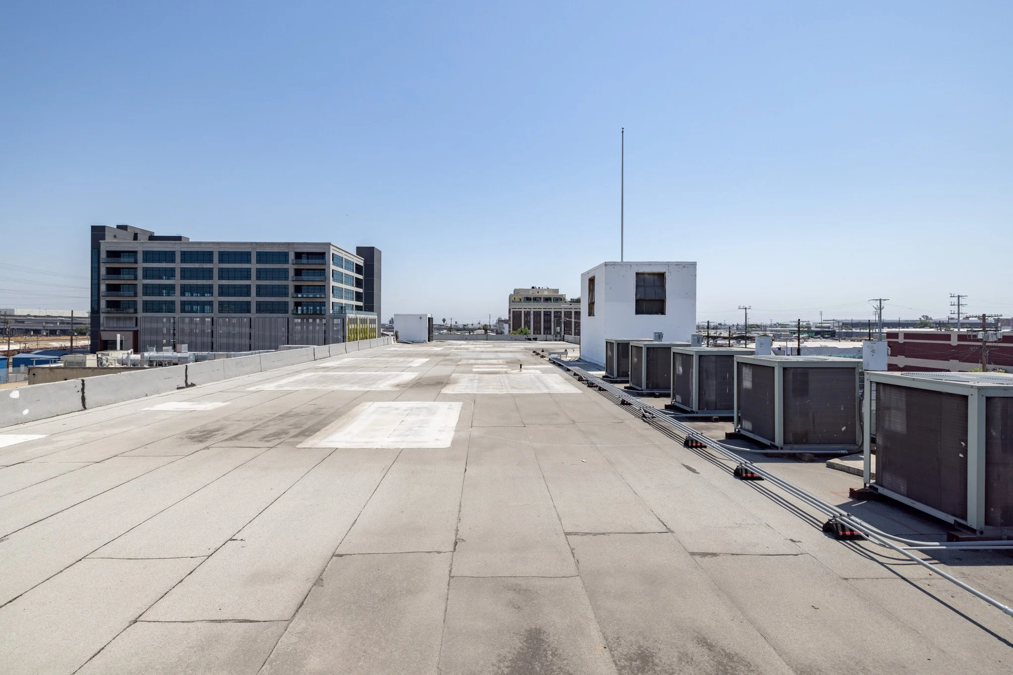 Wide view of a flat rooftop with HVAC units, a small white structure, and a modern office building in the background under a clear blue sky.