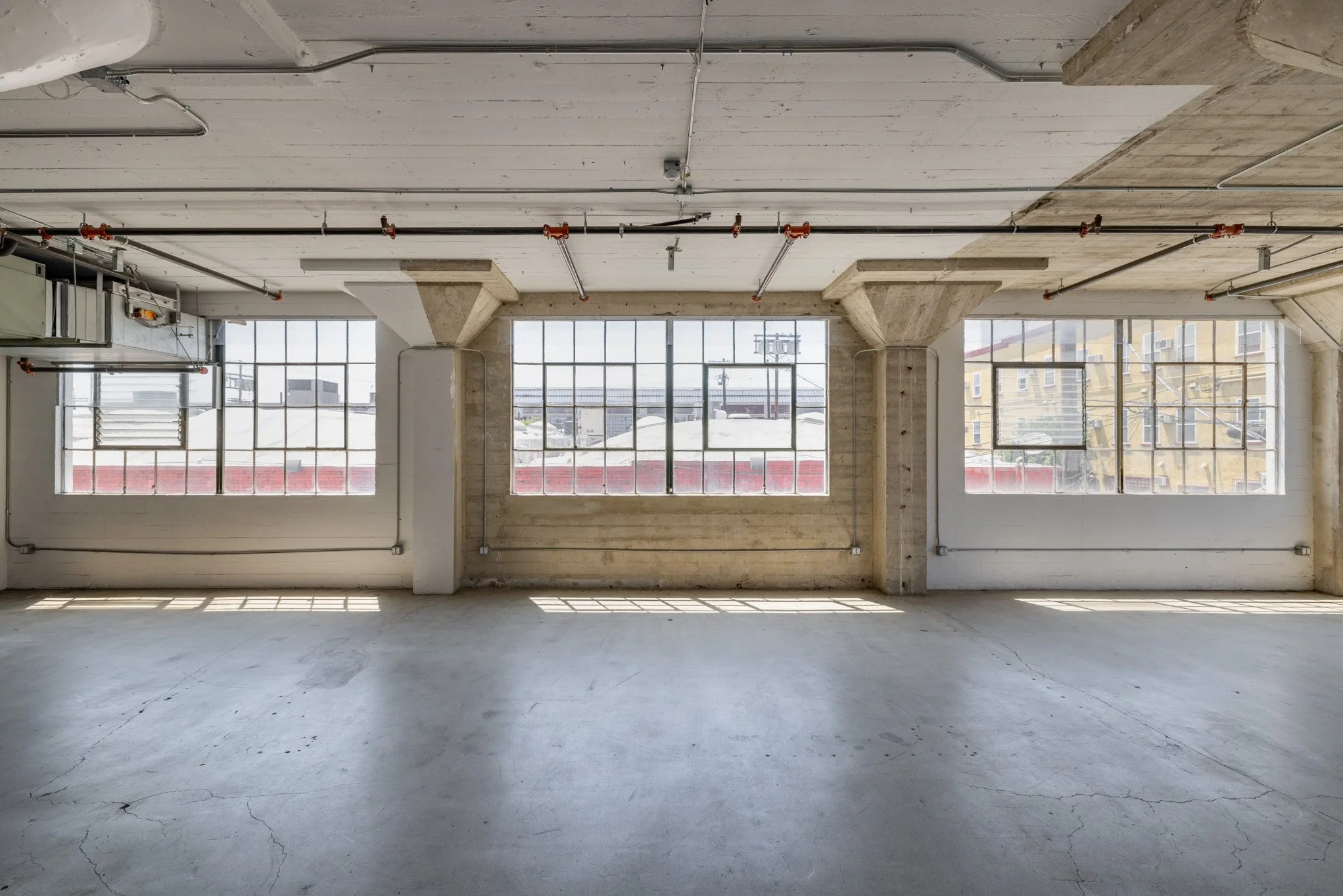 Empty industrial space with three large windows, concrete floor, exposed ceiling pipes, and concrete columns.