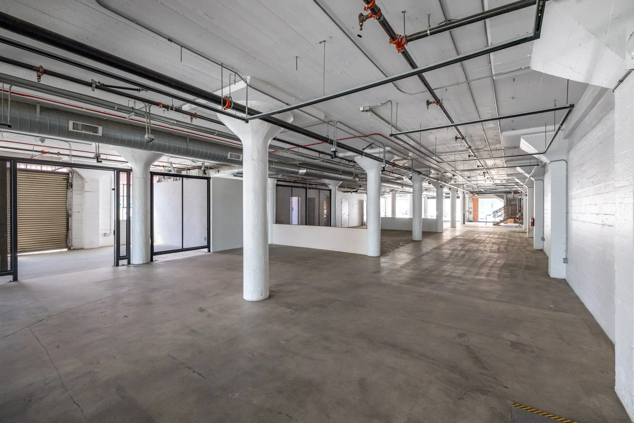 Empty industrial or commercial building with concrete floor, white walls, and exposed pipes and ducts on the ceiling.