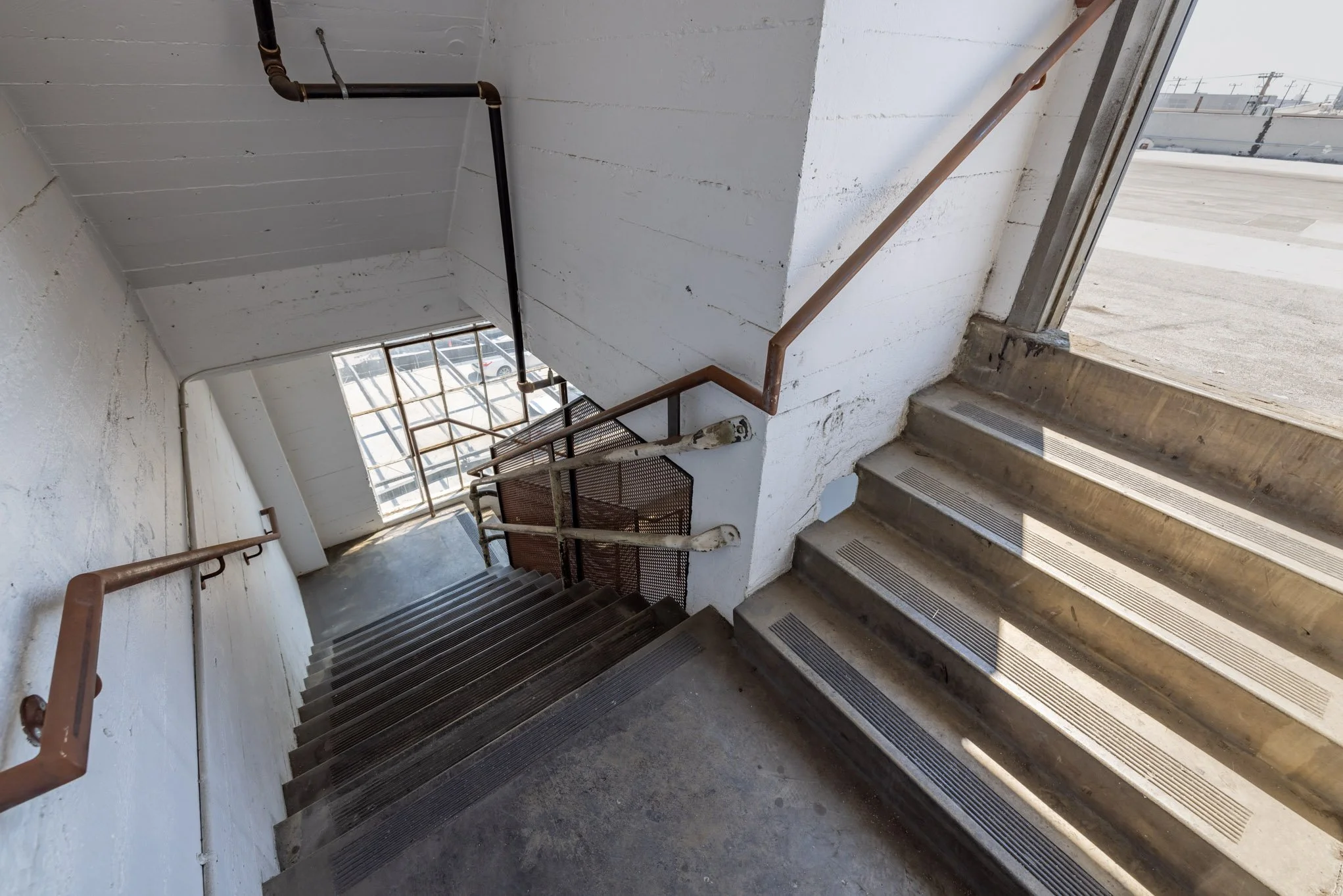 Indoor metal staircase with cobwebs, brown handrails, and vented steps, leading down to a lower level near a window with a parking lot view.