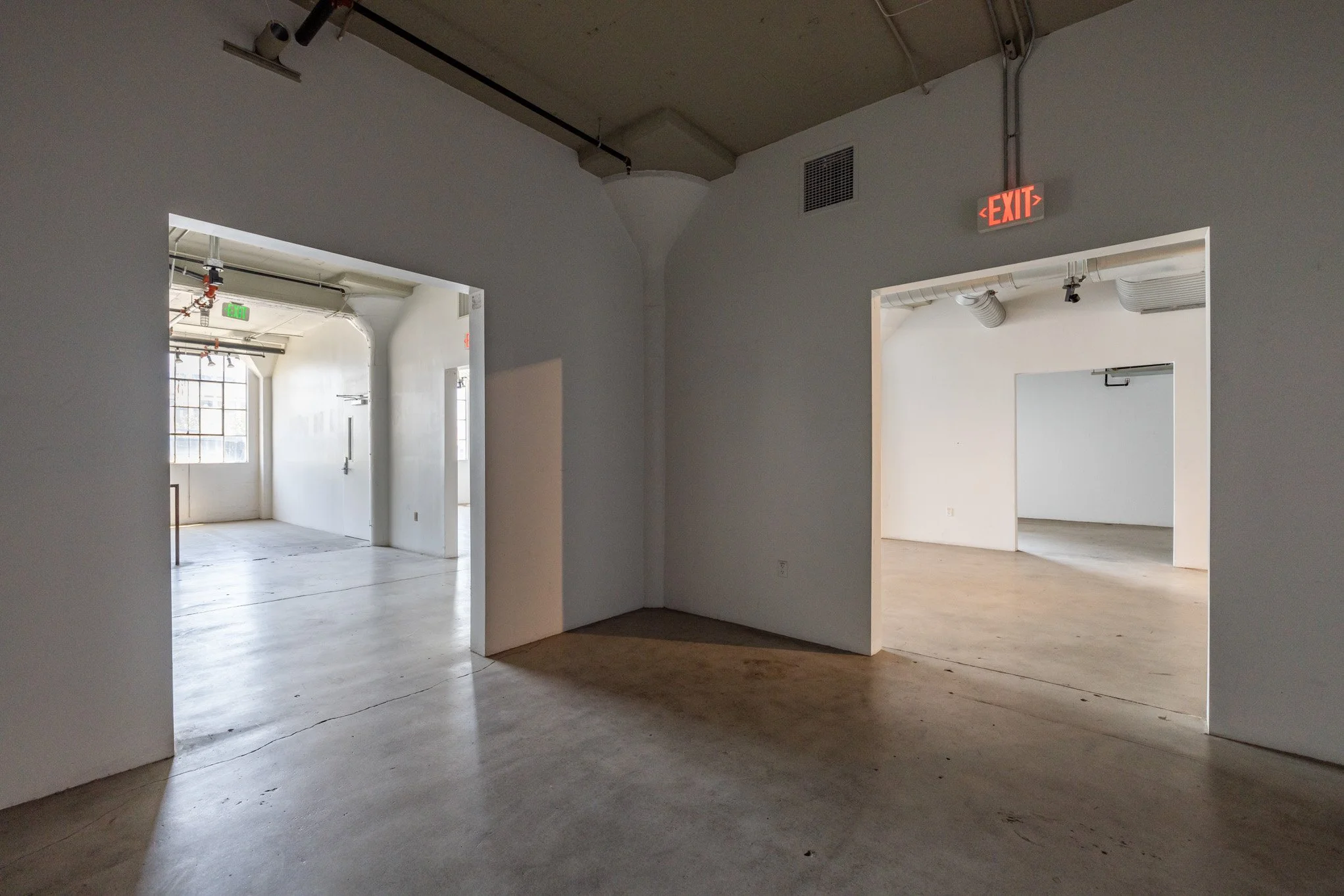 Interior of an empty industrial-style building with white walls and open doorways, visible ceilings with exposed pipes, and an exit sign.