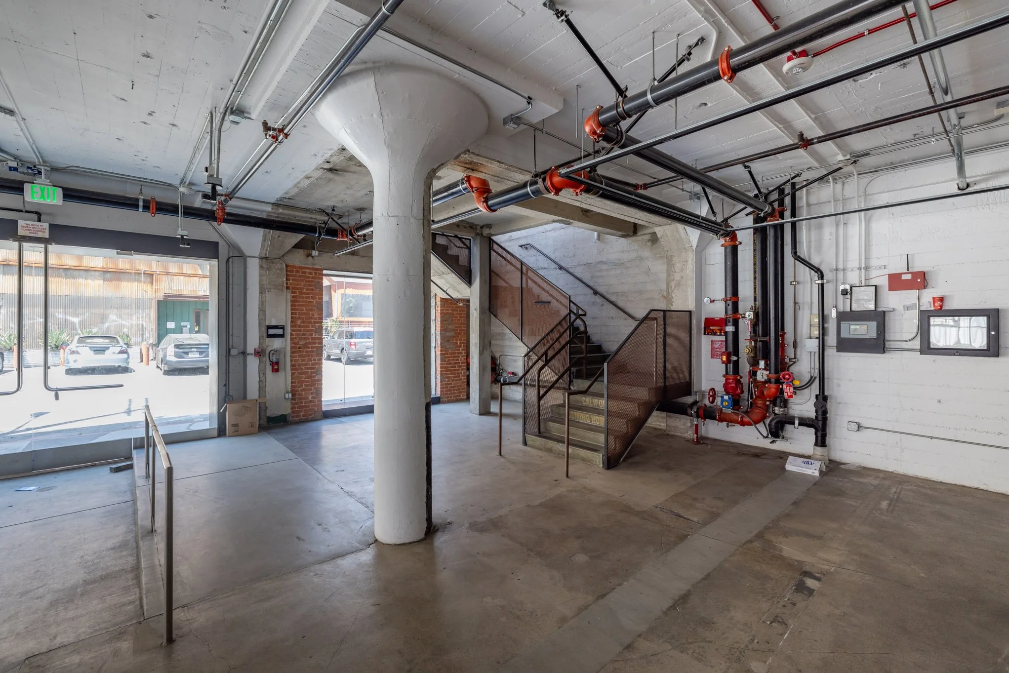 Inside view of an industrial building with exposed pipes, a concrete staircase with metal railing, and a brick wall near large glass windows showing parked cars outside.