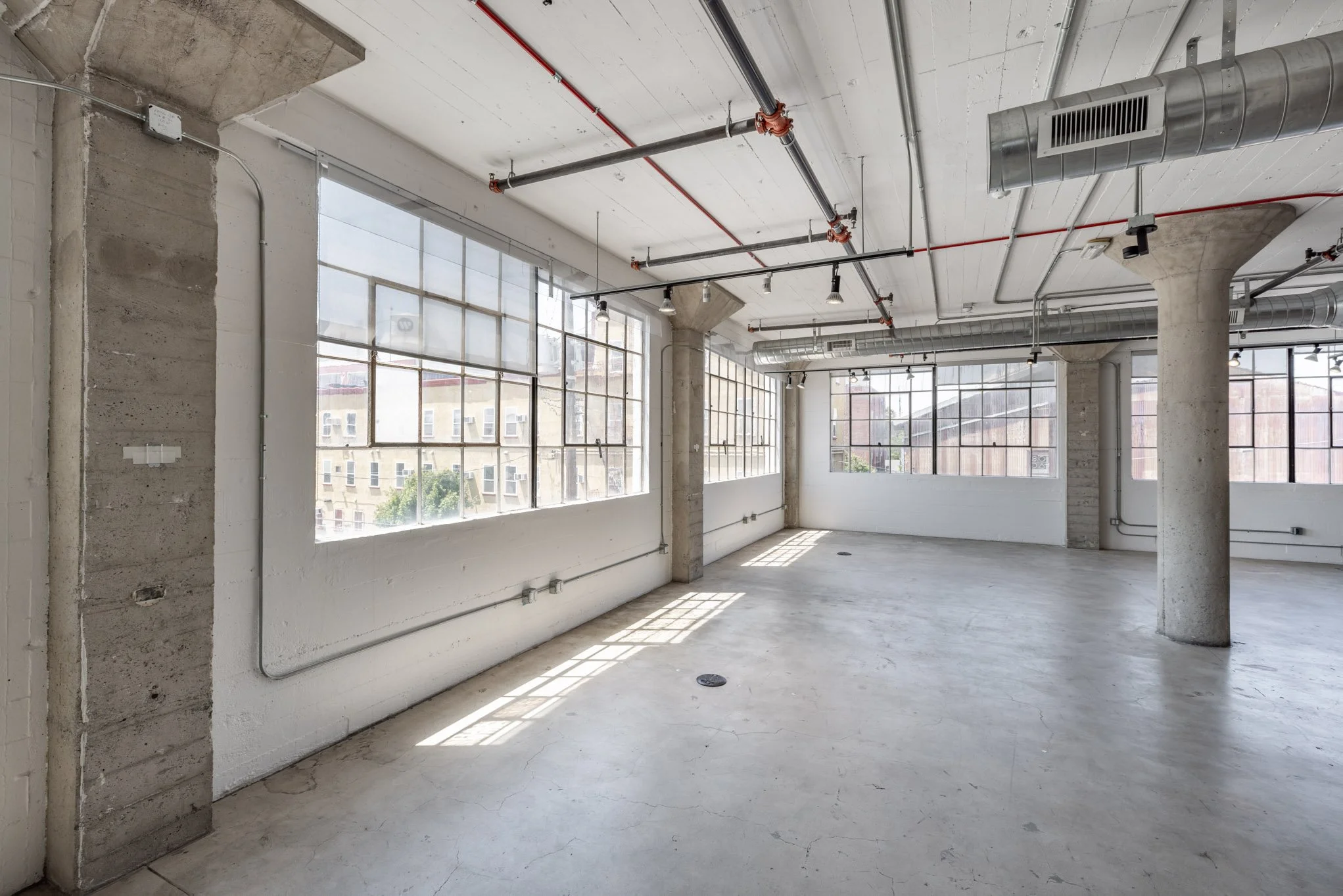 Empty industrial-style office space with large windows, exposed ductwork, and concrete pillars.
