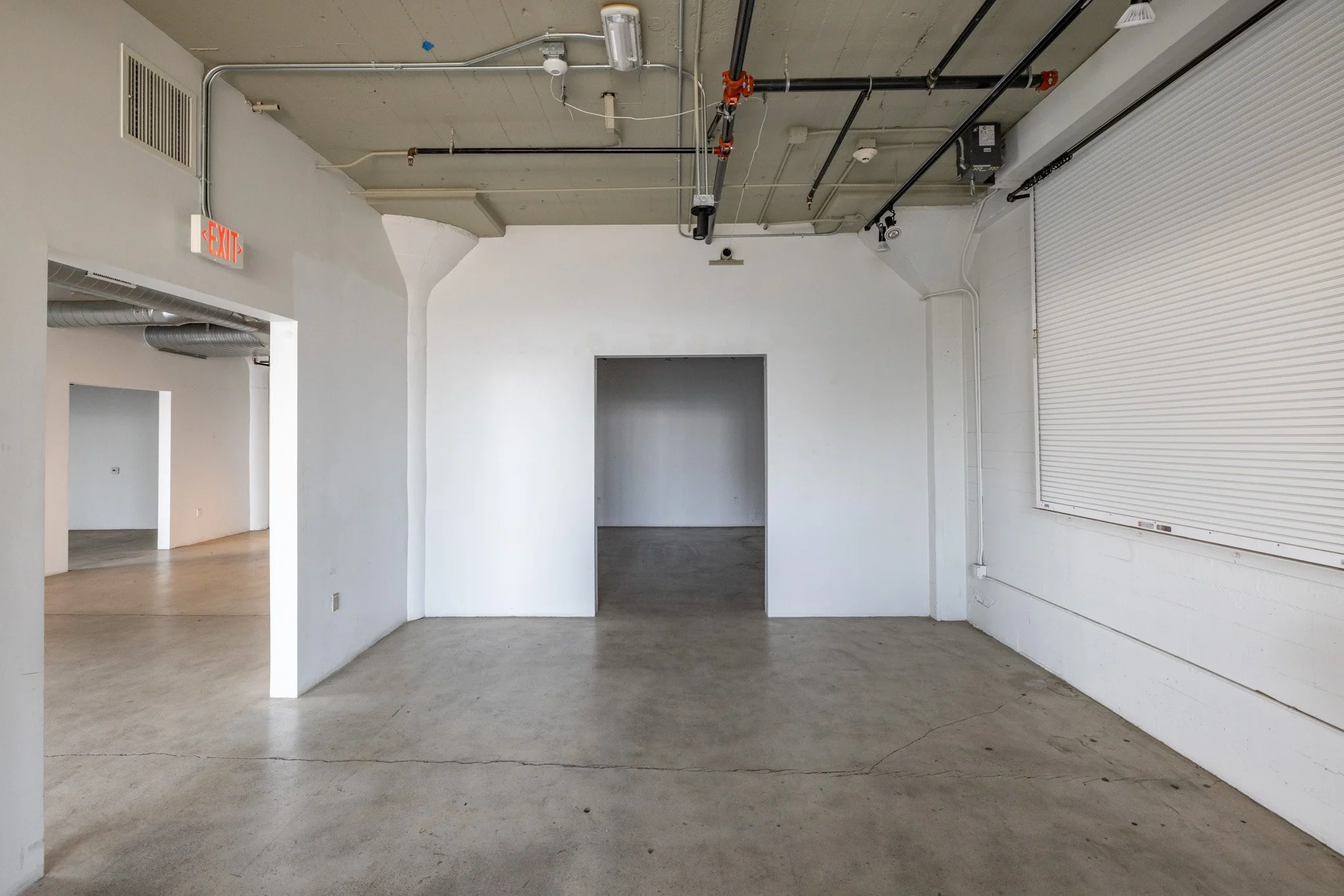 Empty industrial space with exposed ceiling pipes, concrete floor, white walls, and an exit door with a red "EXIT" sign.