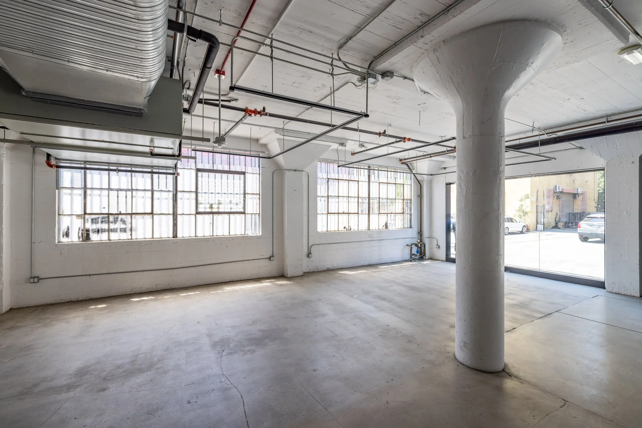 Empty commercial or industrial space with large windows, concrete floor, exposed pipes, and a support column.
