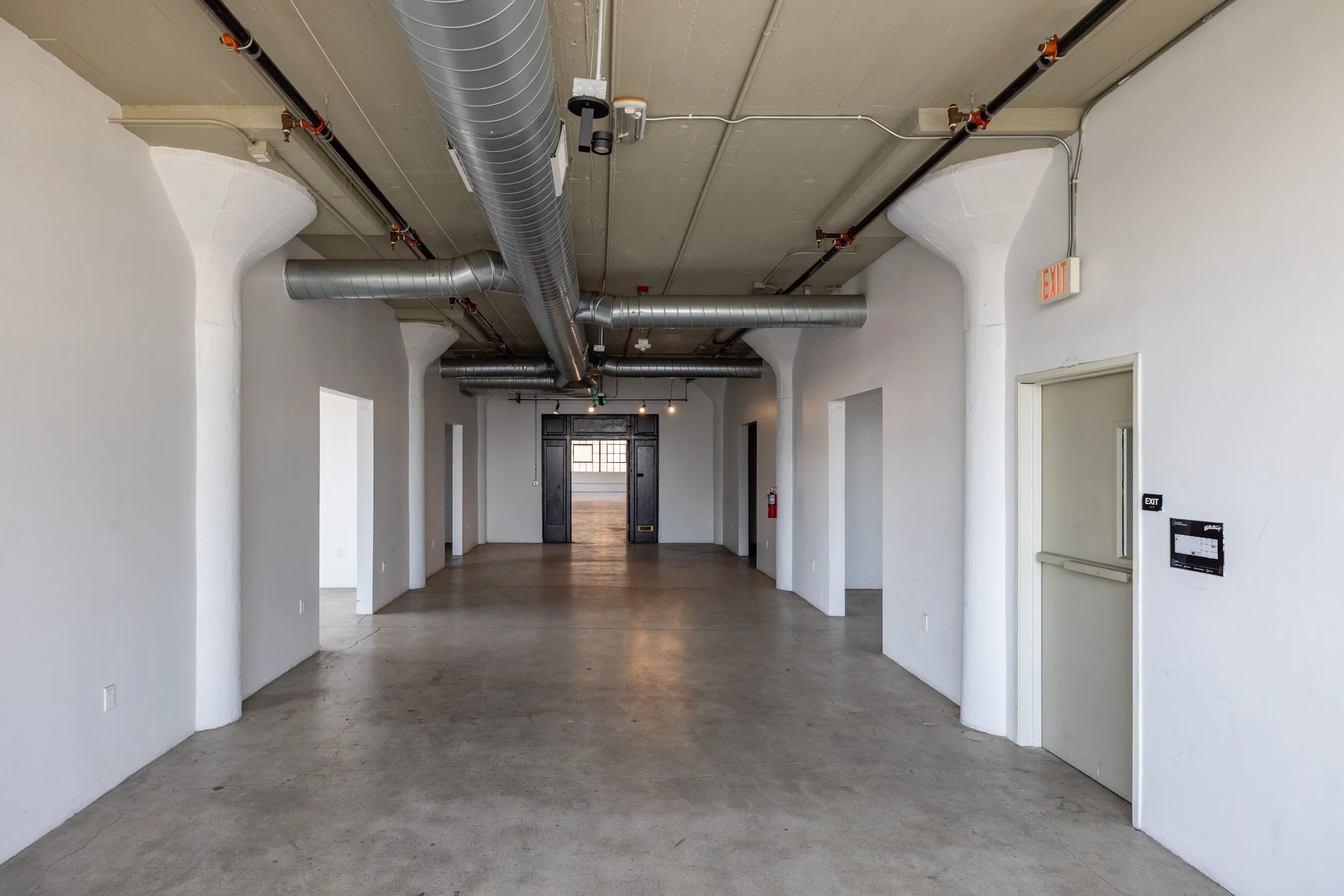 Empty interior space with white walls, concrete floor, exposed metal ductwork on ceiling, doorway at the back, and fire safety equipment on the wall.