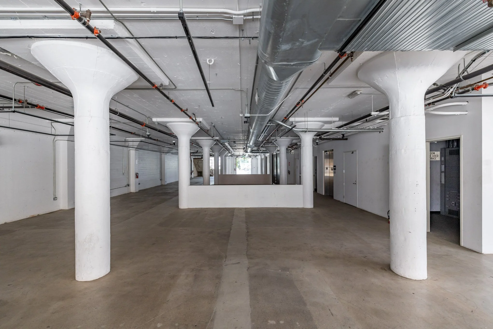Empty industrial or commercial underground parking garage with white support columns, exposed piping and ducts on the ceiling, and concrete flooring.