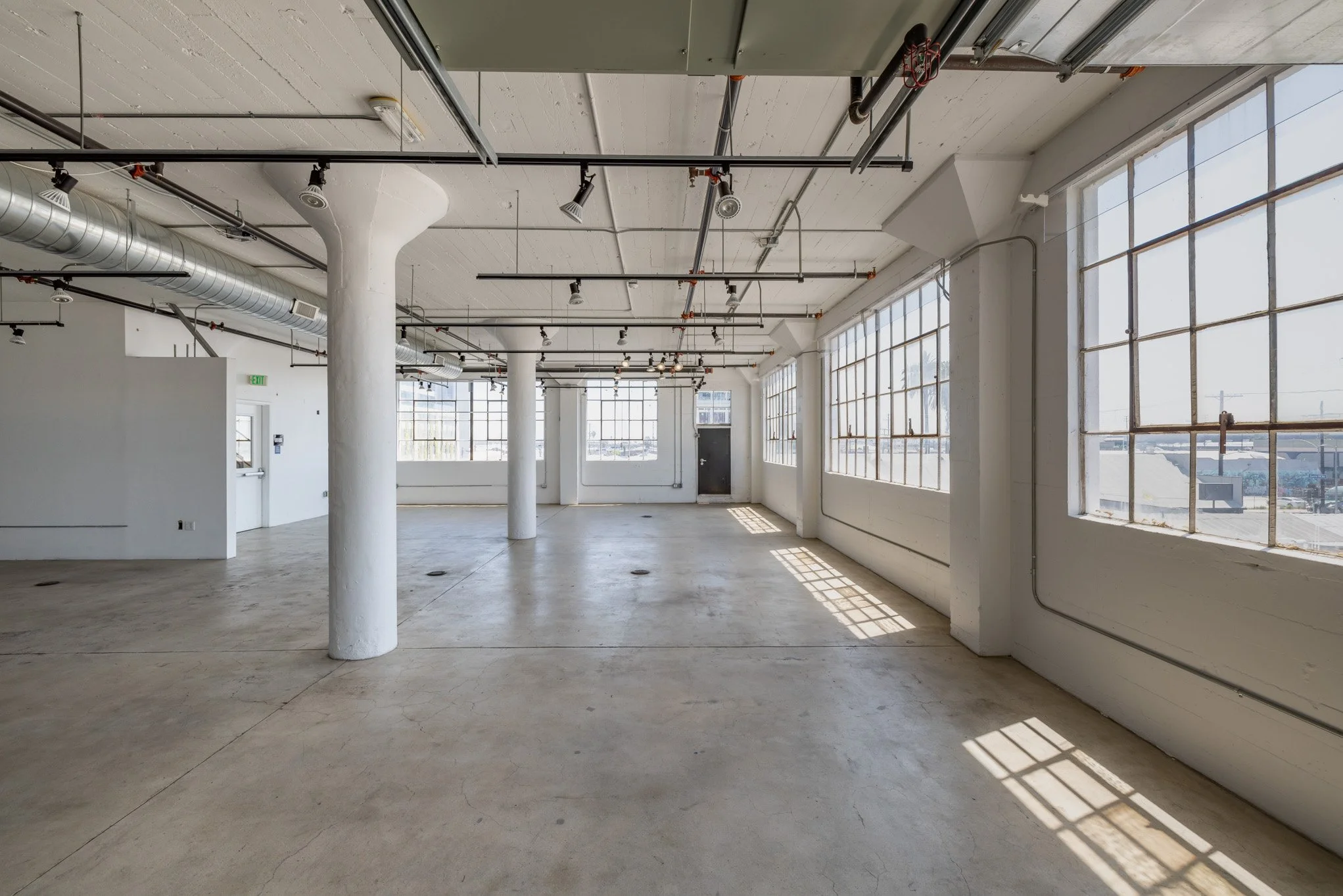 Empty industrial-style loft with large windows, concrete floor, exposed pipes and columns, and sunlight streaming in.
