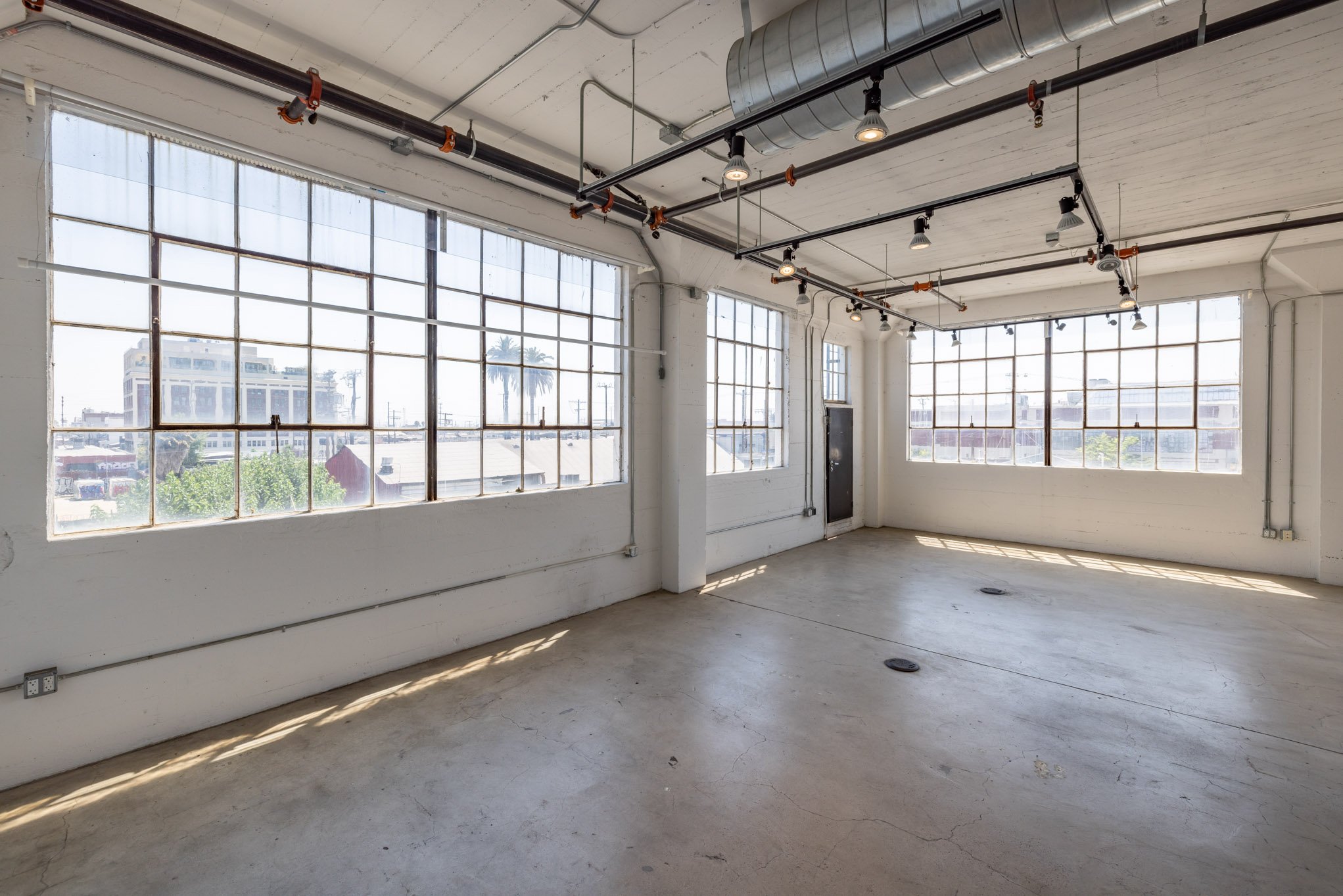 Empty industrial-style room with large grid windows, concrete floor, exposed ceiling pipes, and track lighting, with sunlight streaming in.
