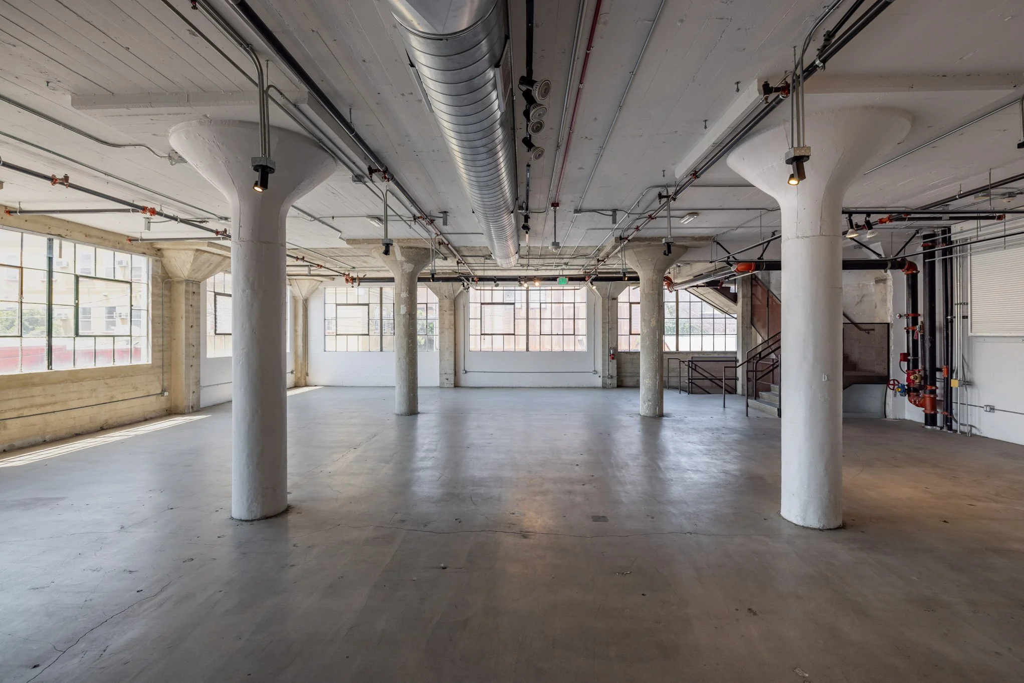 Empty industrial-style interior space with concrete floors, large windows, and white pillars, featuring exposed ductwork and piping.
