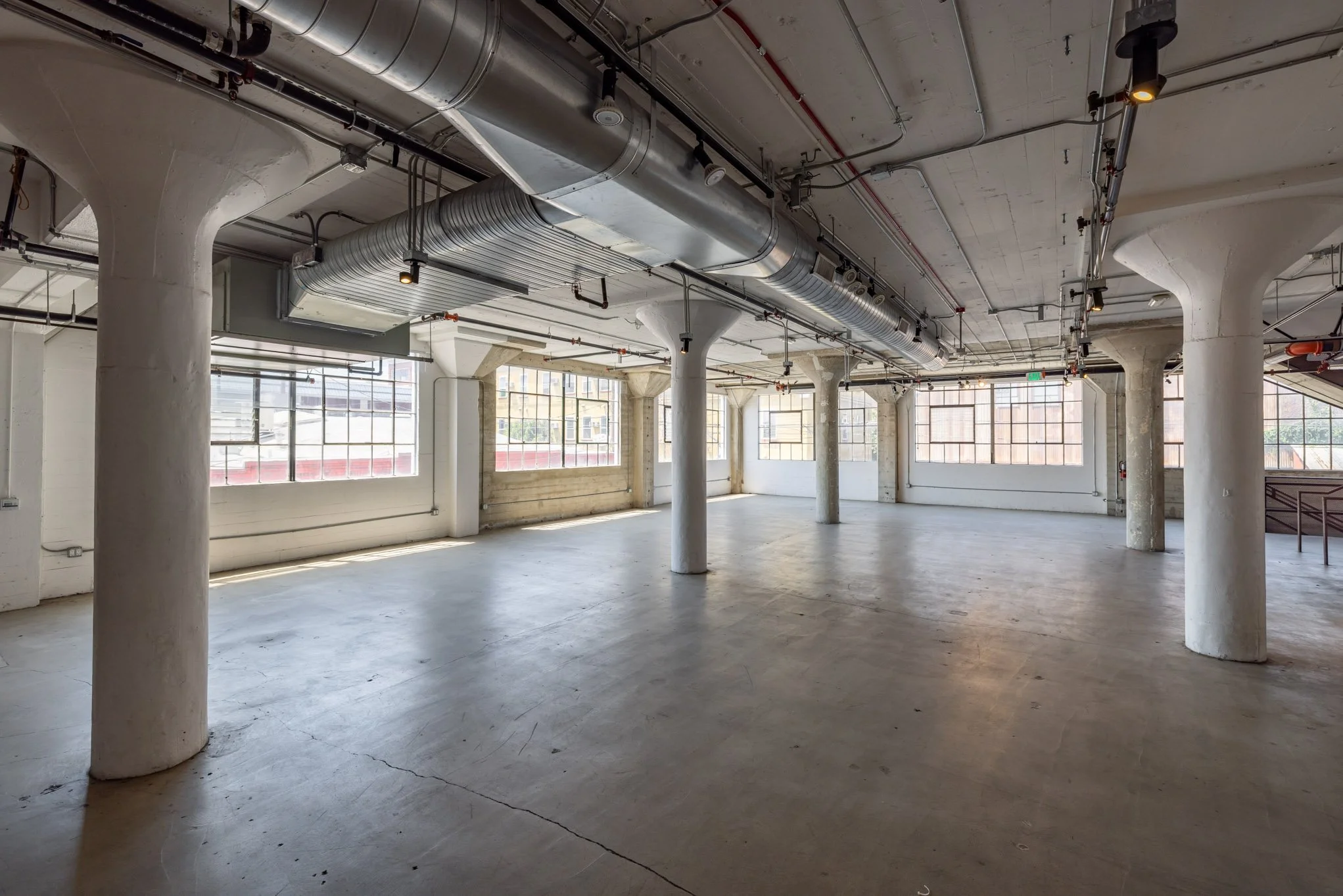 Empty industrial-style room with large windows, concrete floors, ceiling pipes, and white columns.