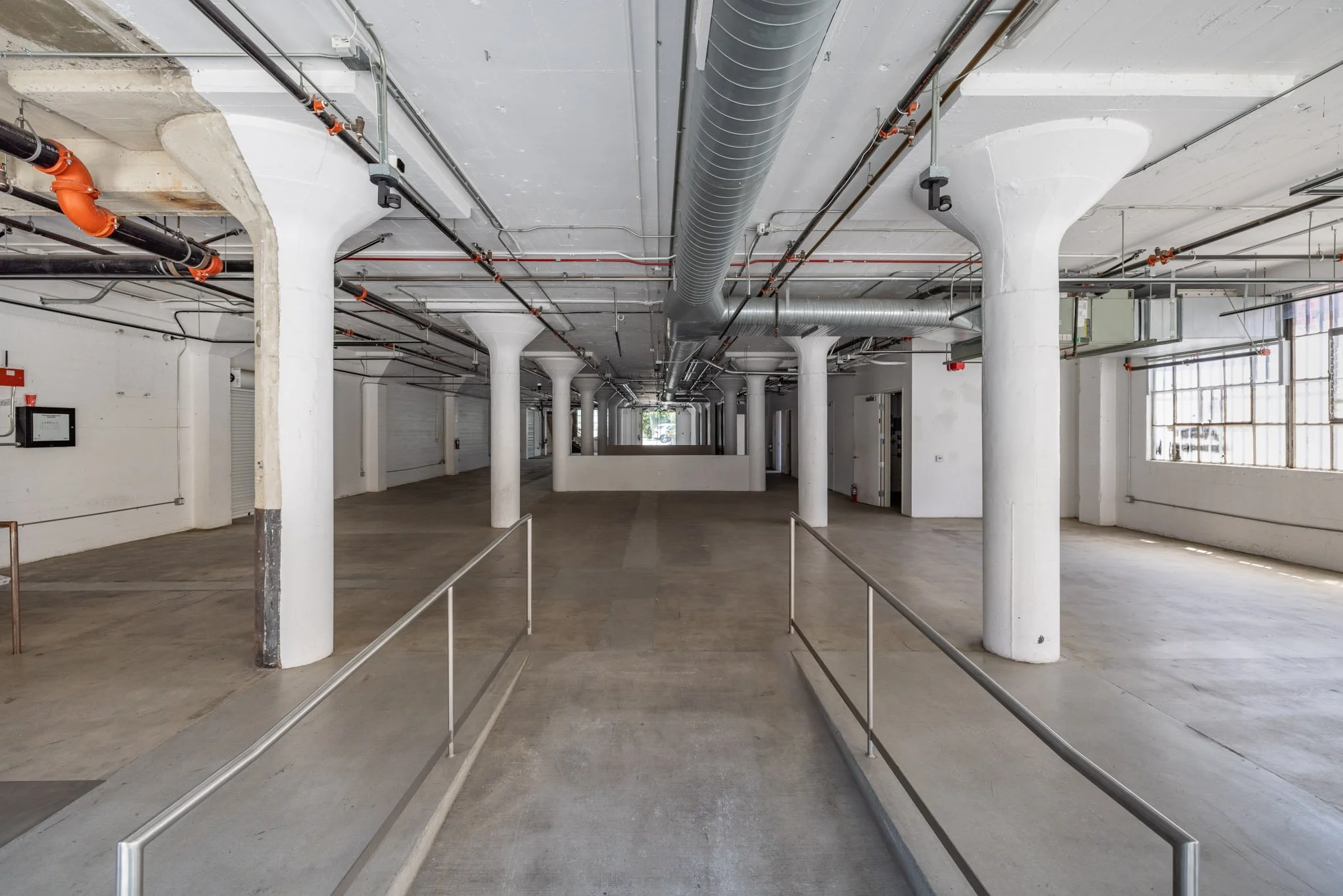 Empty industrial or commercial space with white support columns, exposed pipes on the ceiling, large windows on the right, and a wheelchair accessible ramp.