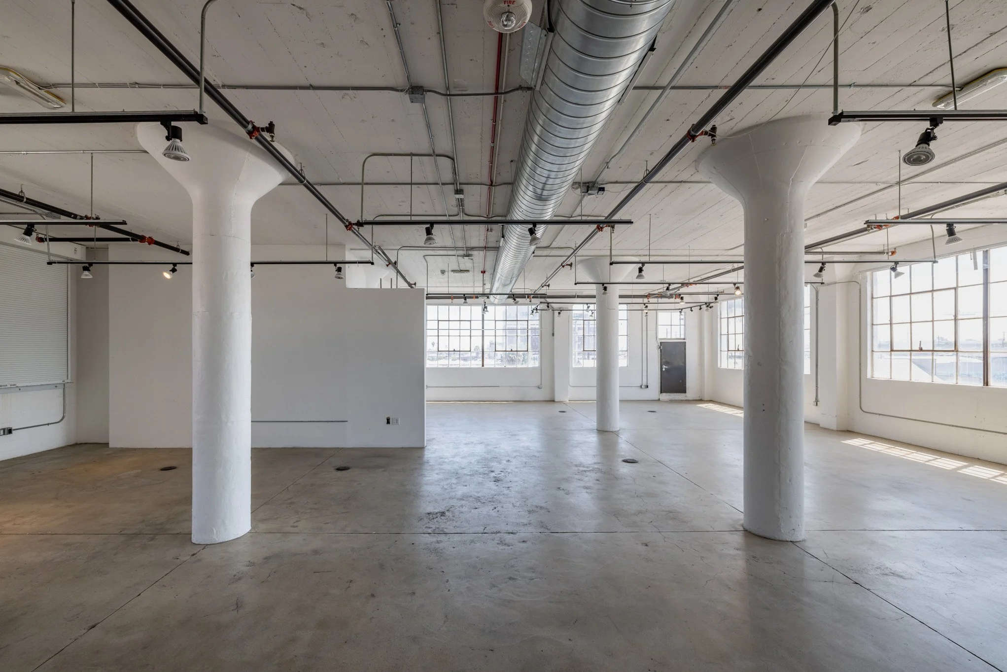 Empty industrial-style loft with white walls, large windows, and exposed ductwork and pipes on the ceiling.