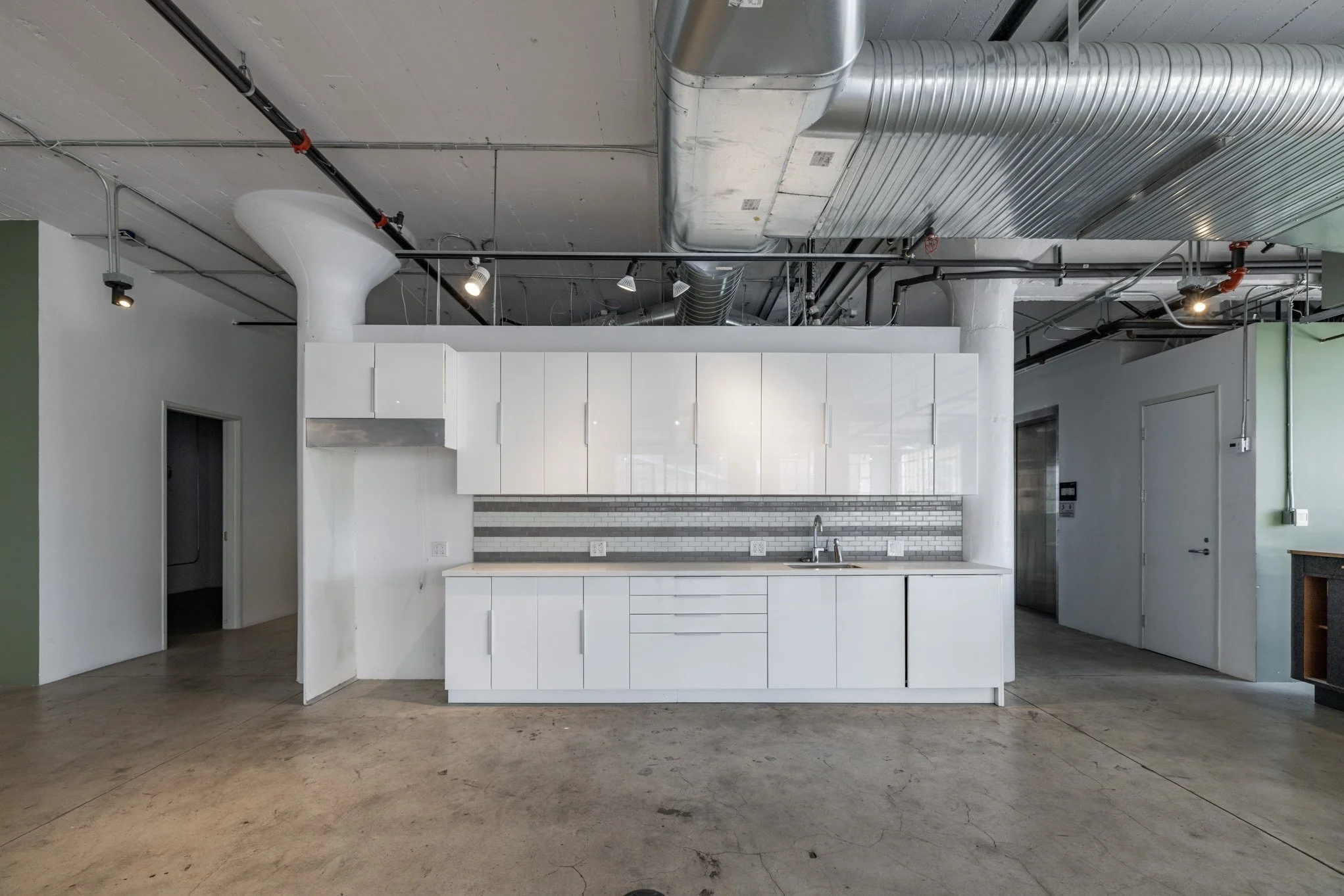 Empty modern kitchen with white cabinets, gray backsplash, stainless steel vent hood, industrial ceiling with exposed ductwork, and concrete floor.
