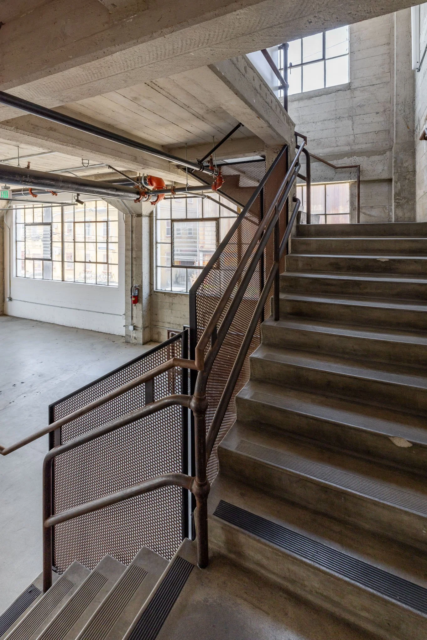 Interior view of a concrete staircase in an industrial-style building with large windows and exposed piping.