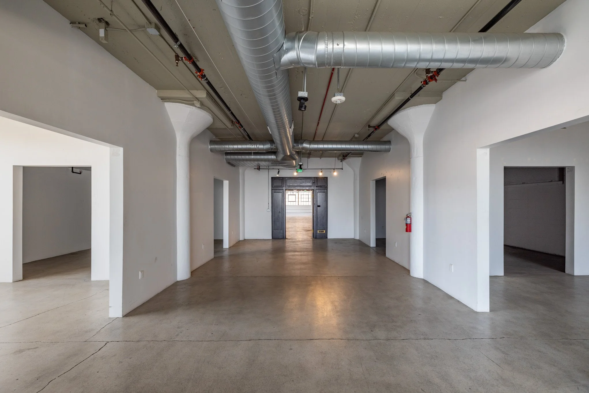 Empty commercial space with white walls, polished concrete floors, large open entrances, an industrial ceiling with visible ductwork, and a fire extinguisher mounted on the right wall.