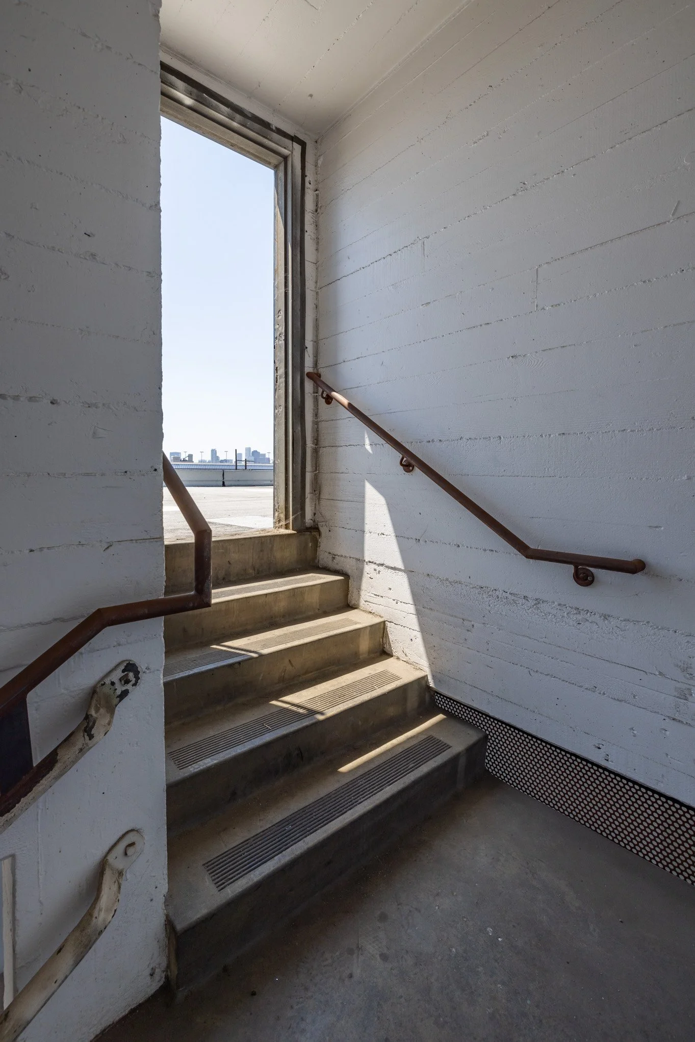 Concrete stairs with a bent handrail next to an open exit door leading to an outdoor rooftop area with city skyline in the distance.