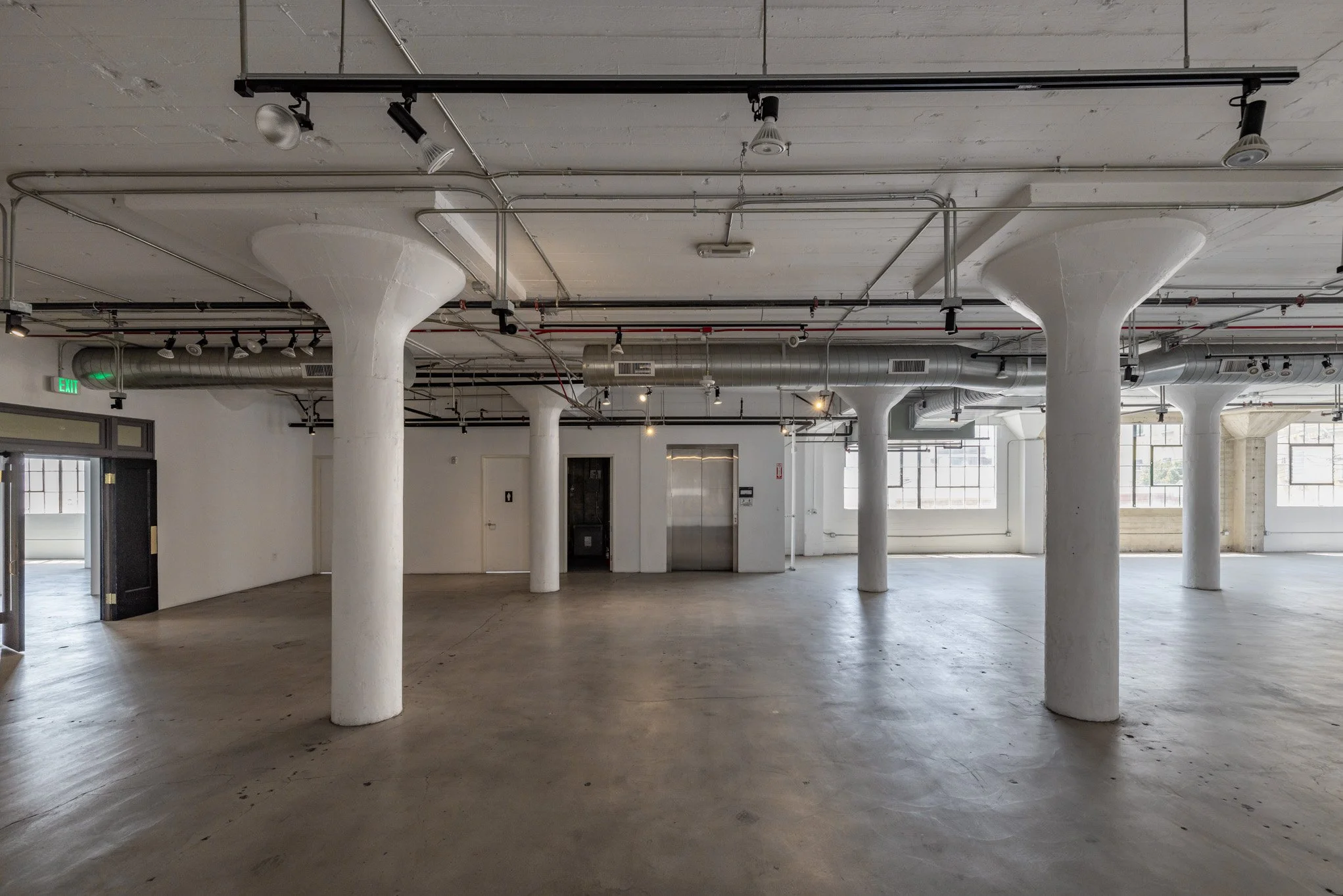 Empty industrial-style room with white pillars, exposed ductwork, large windows, and concrete floors.