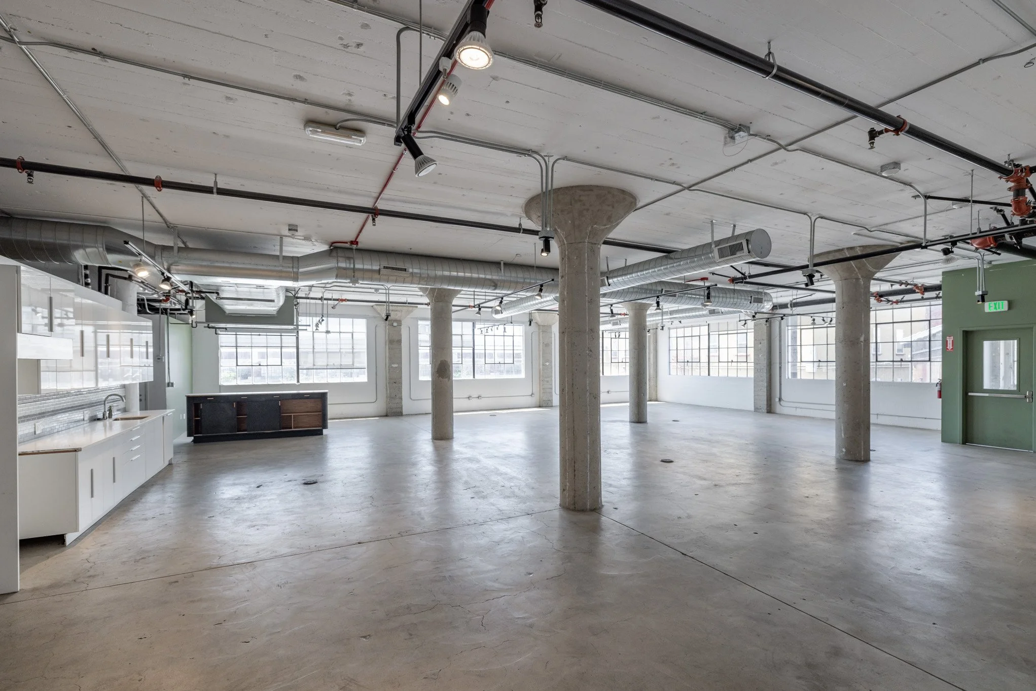 Empty industrial-style room with large windows, concrete floors, exposed ducts, and several concrete support columns.