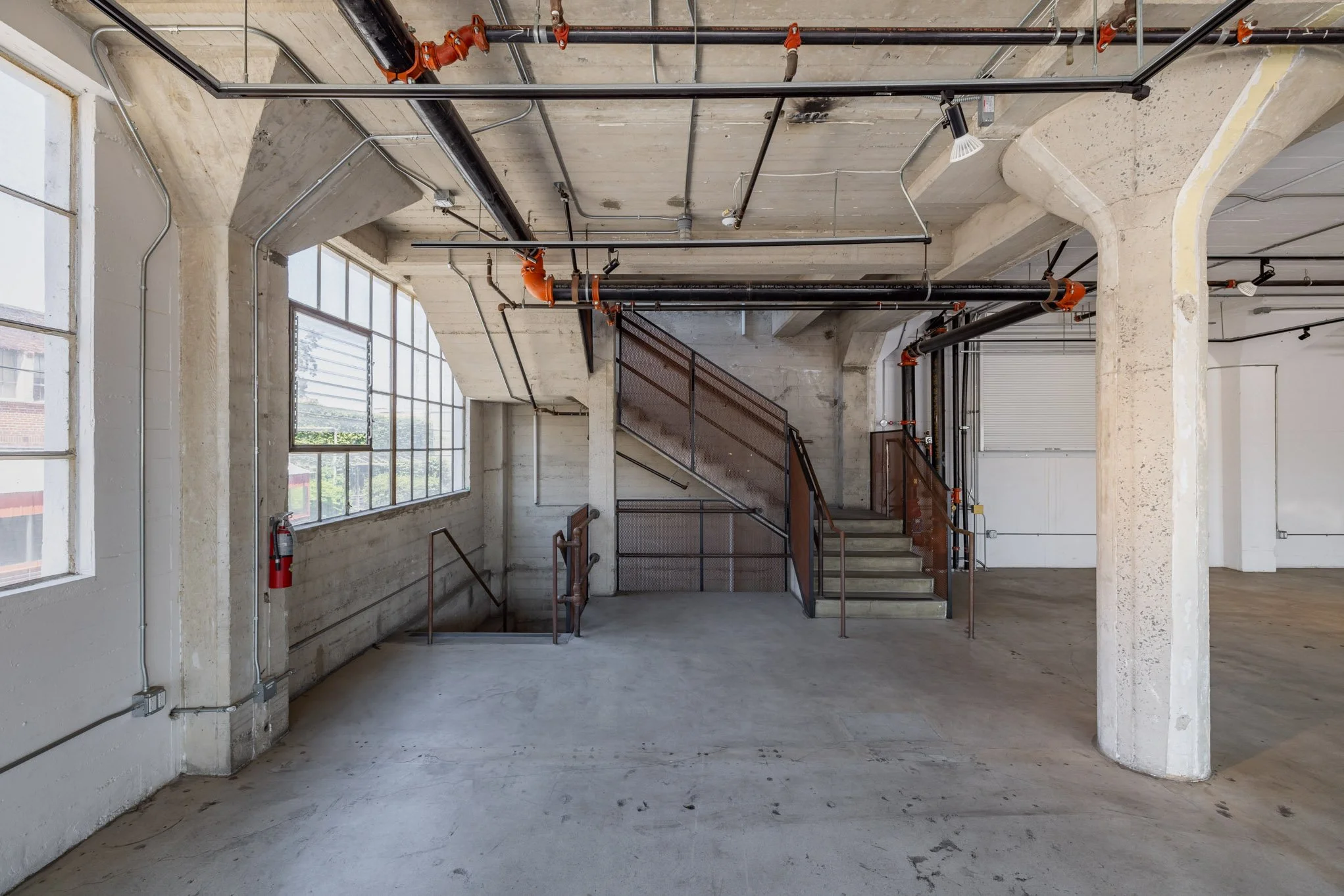 Interior of an industrial-style building with exposed concrete walls, large windows, metal pipes on the ceiling, a staircase with metal railings, and a fire extinguisher mounted on the wall.