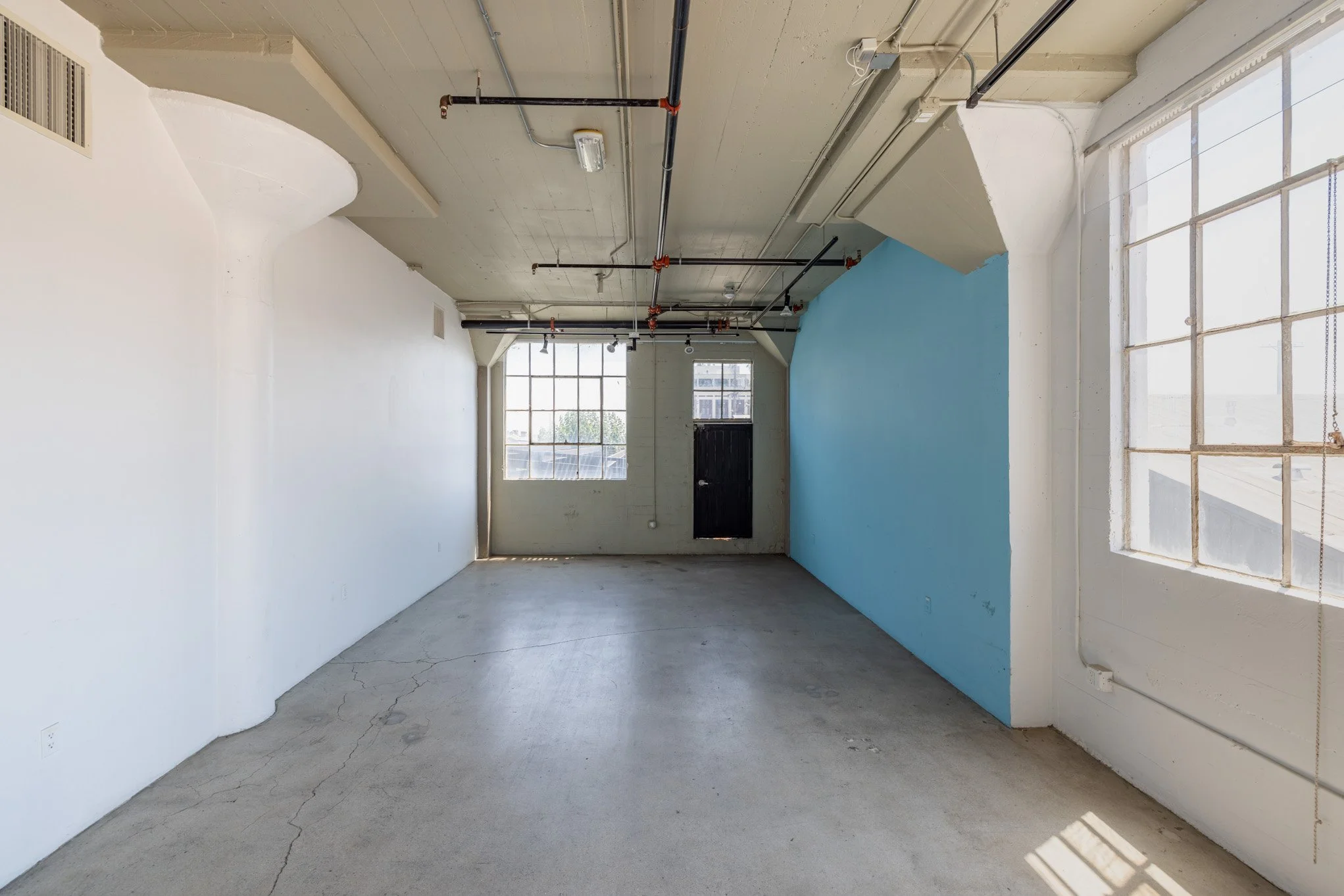 Empty room with white and light blue walls, large windows on the sides, concrete floor with cracks, and ceiling with exposed pipes and electrical conduit.