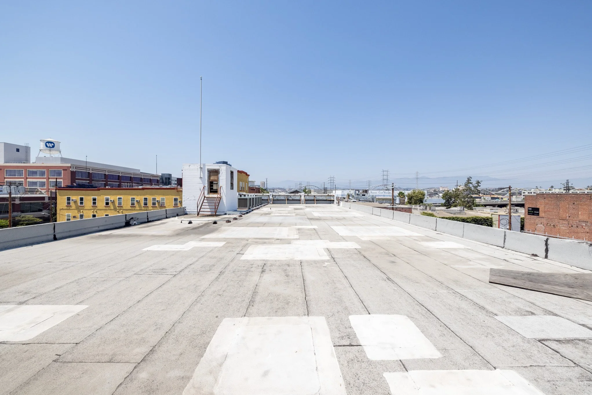 Empty rooftop parking lot with painted white arrow marking and a small white building at the far end, set against a clear blue sky and urban cityscape in the background.
