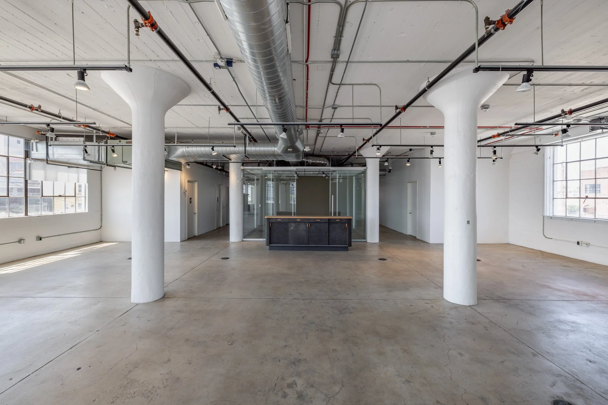 Empty industrial-style office space with concrete floors, white walls, large windows, exposed ductwork, and a small reception desk in the center.