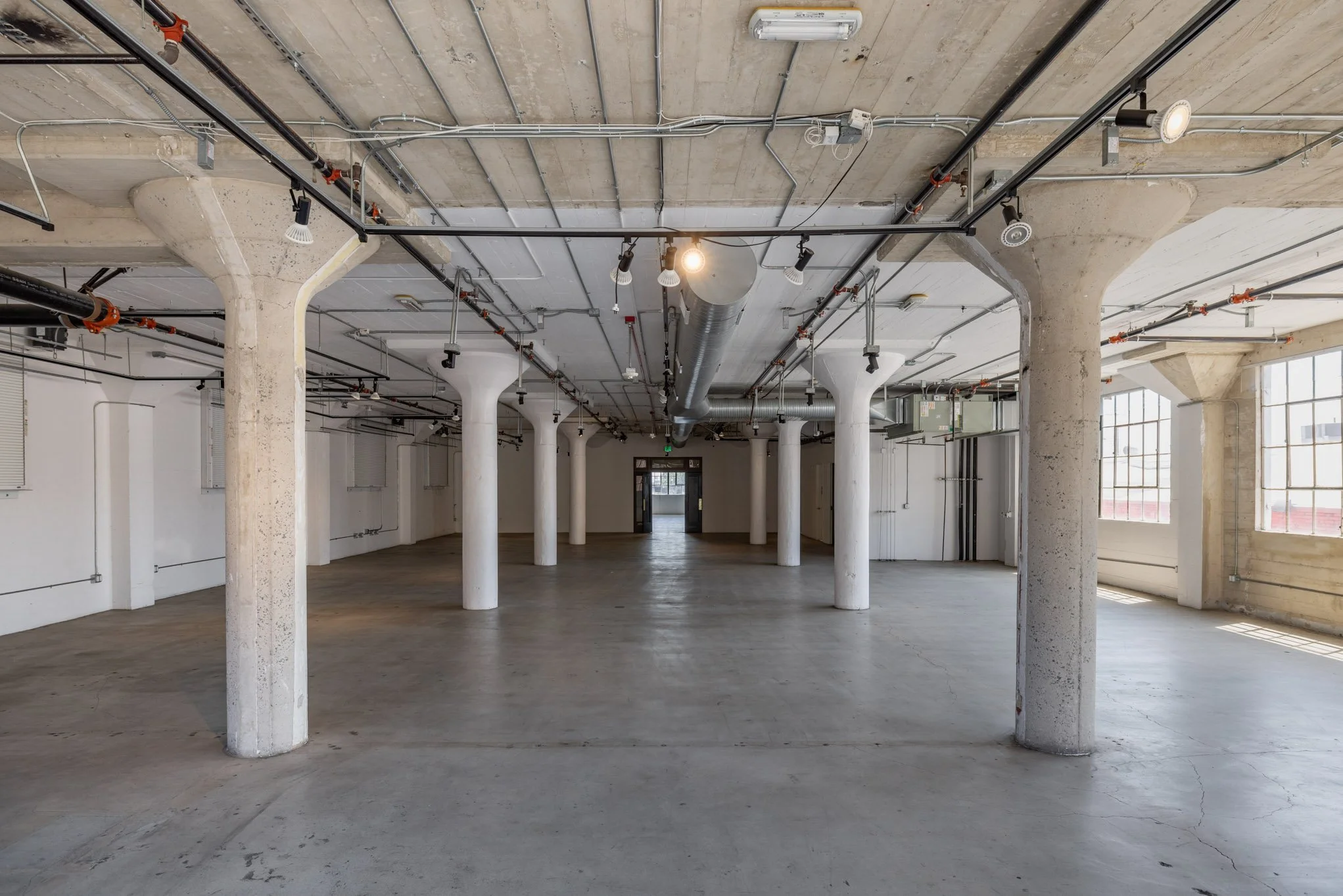 Empty industrial-style interior space with concrete pillars, exposed ceiling piping, and large windows.