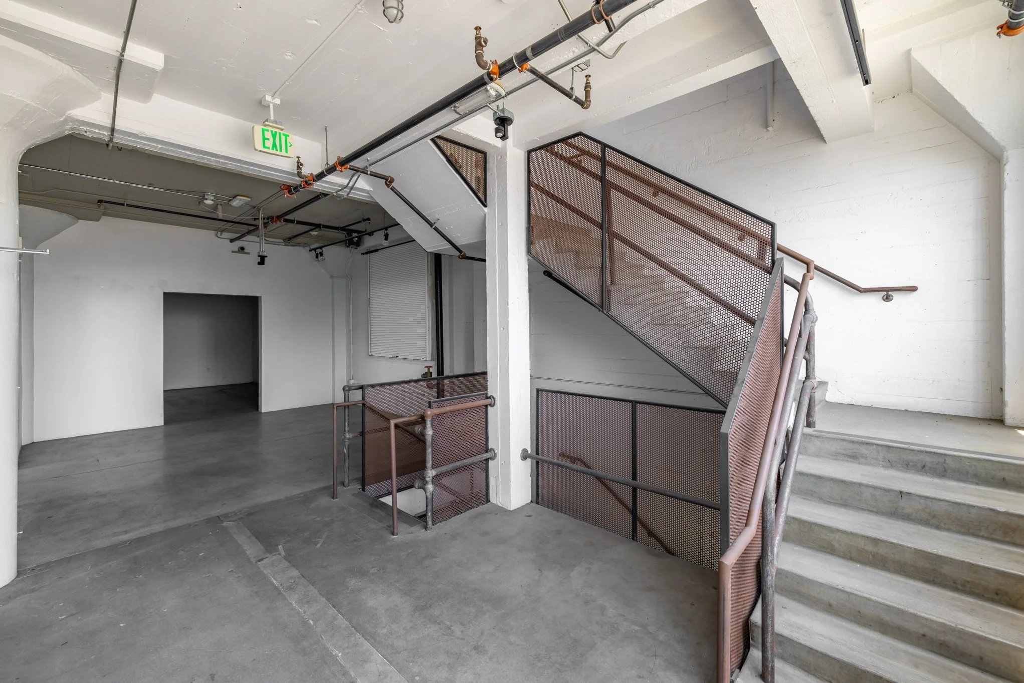 Empty industrial-style staircase and hallway with white-painted brick walls, concrete floors, exposed piping, and an illuminated green exit sign.